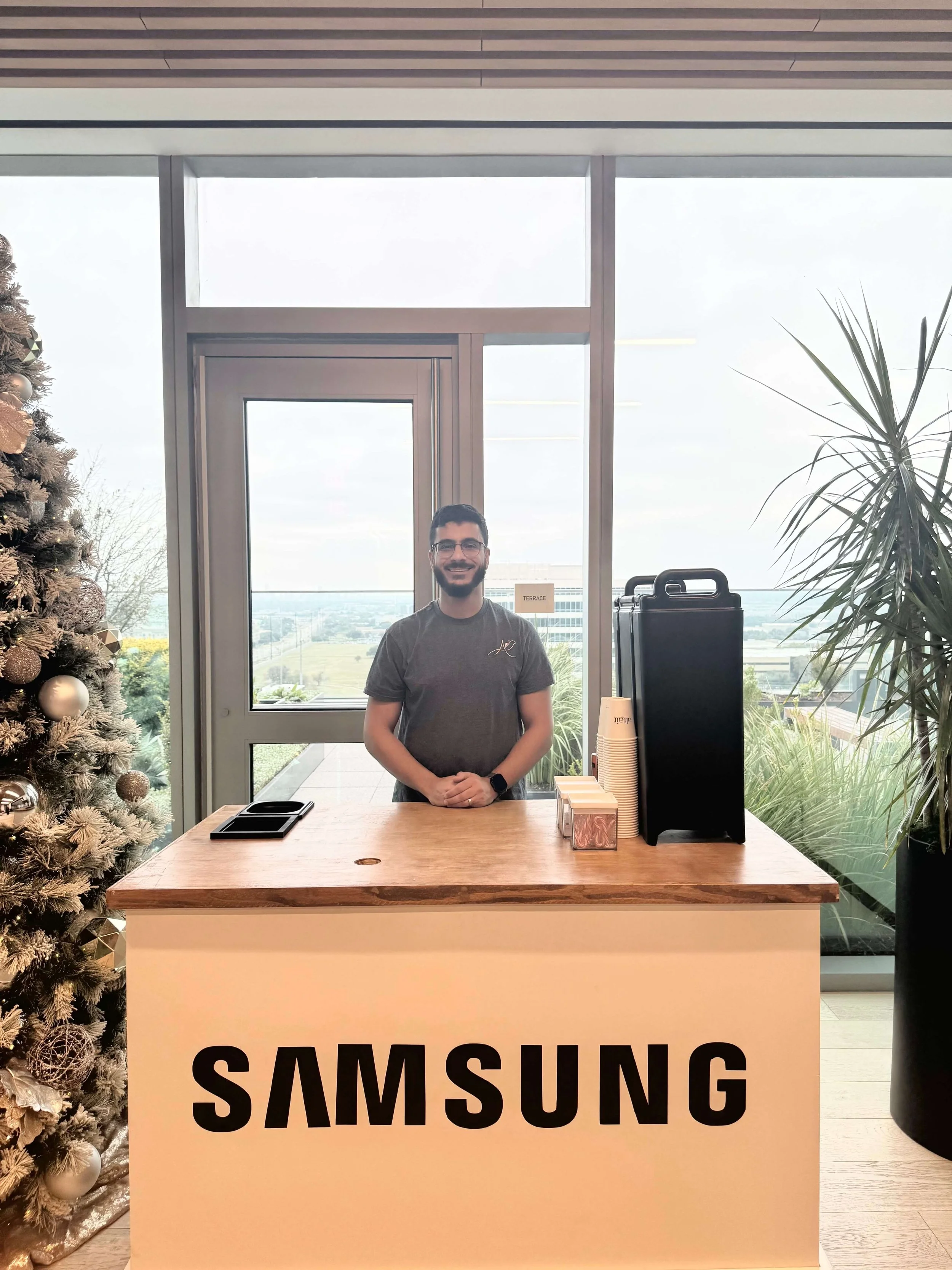 A professional barista standing behind a Samsung custom branded coffee cart counter in a modern office or showroom, with a Christmas tree to the left and large windows in the background showing a view of the outdoors.