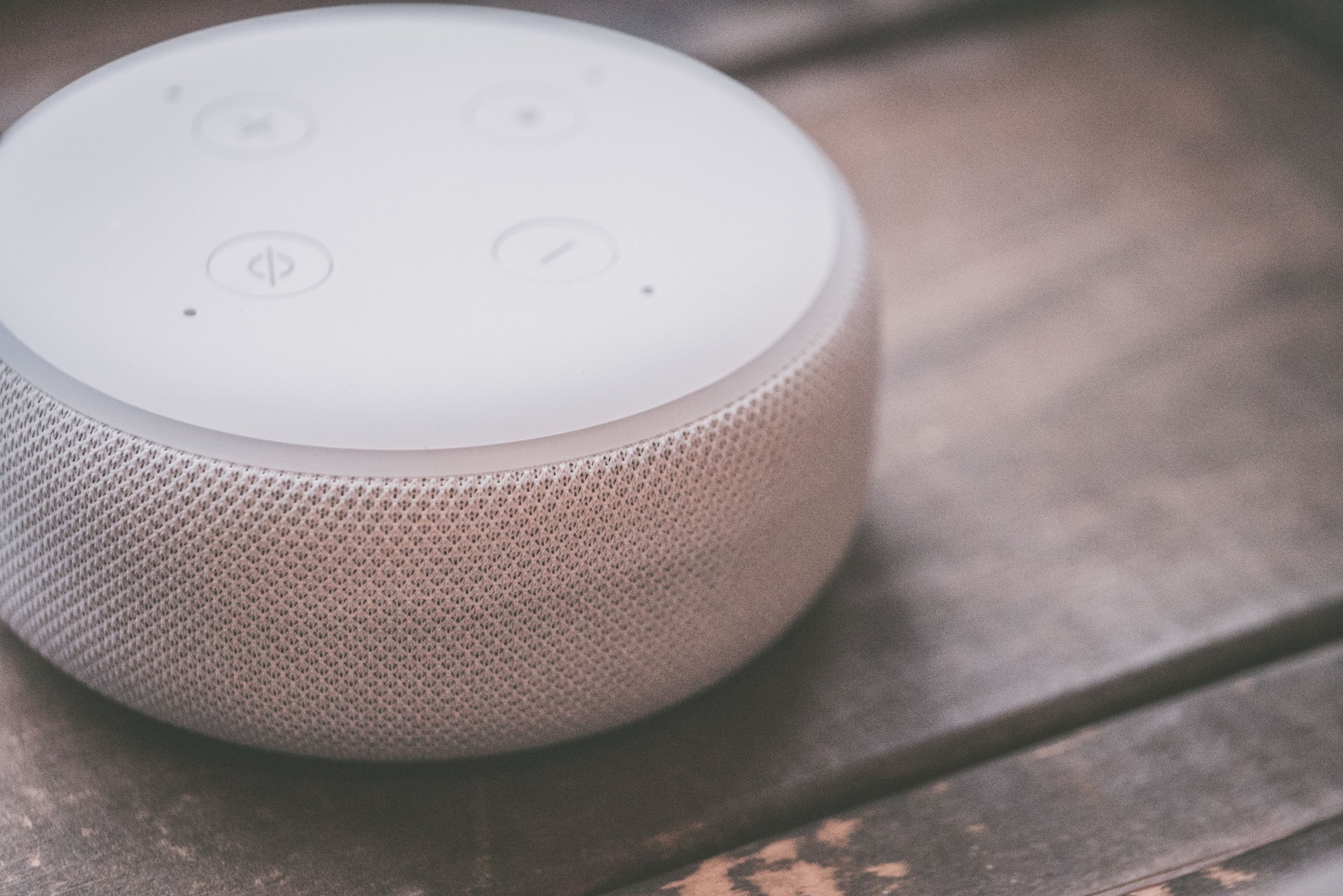 Close-up of an Amazon Echo device with a white top surface and a pinkish textured fabric body, placed on a dark wooden surface.
