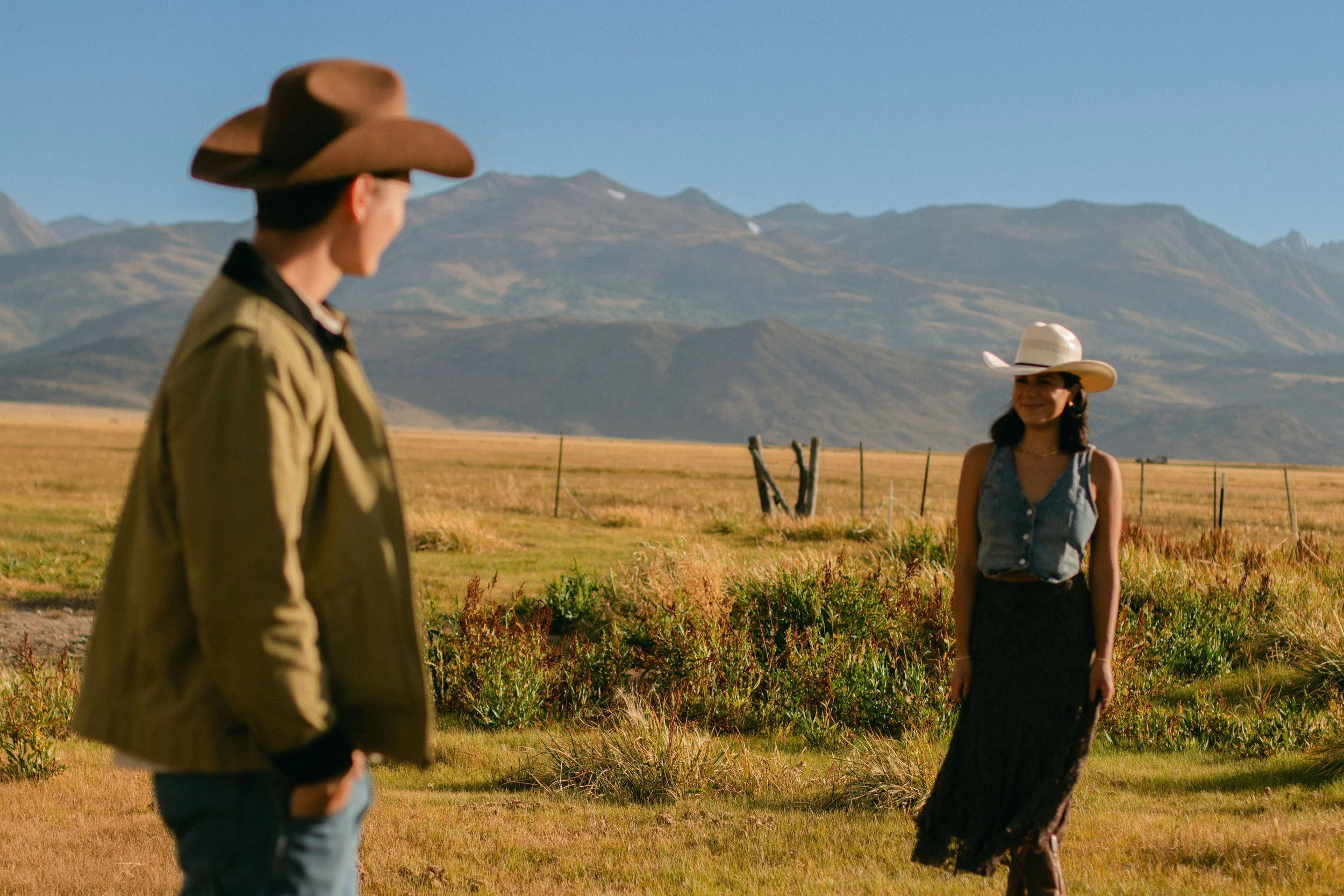 A man and woman standing outdoors in a field with mountains in the background, both wearing cowboy hats.