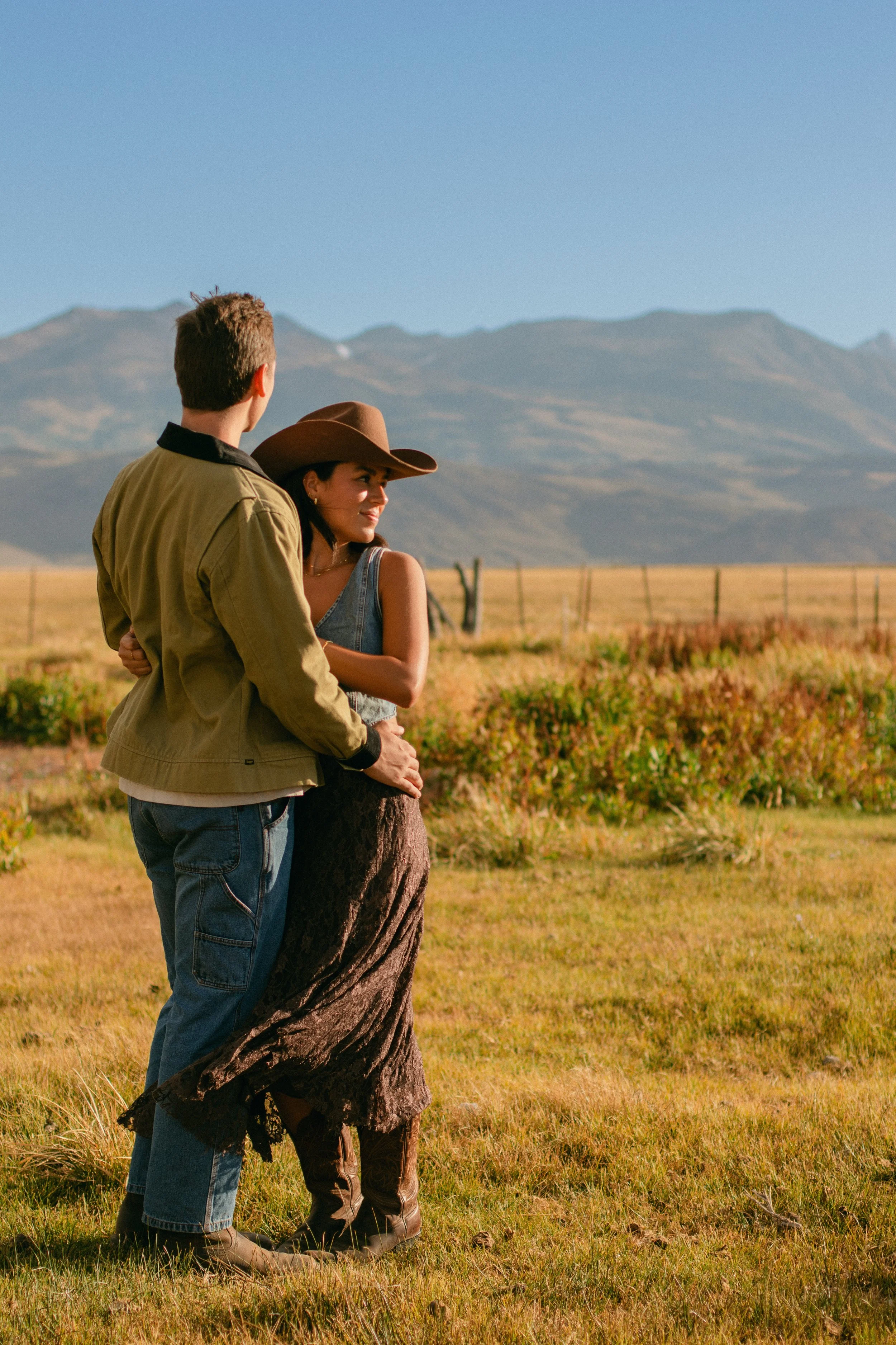 A couple standing close together in a field with mountains in the background, during daytime. The man is wearing a tan jacket and blue jeans, while the woman is dressed in a denim vest, a brown skirt, and a wide-brimmed hat.