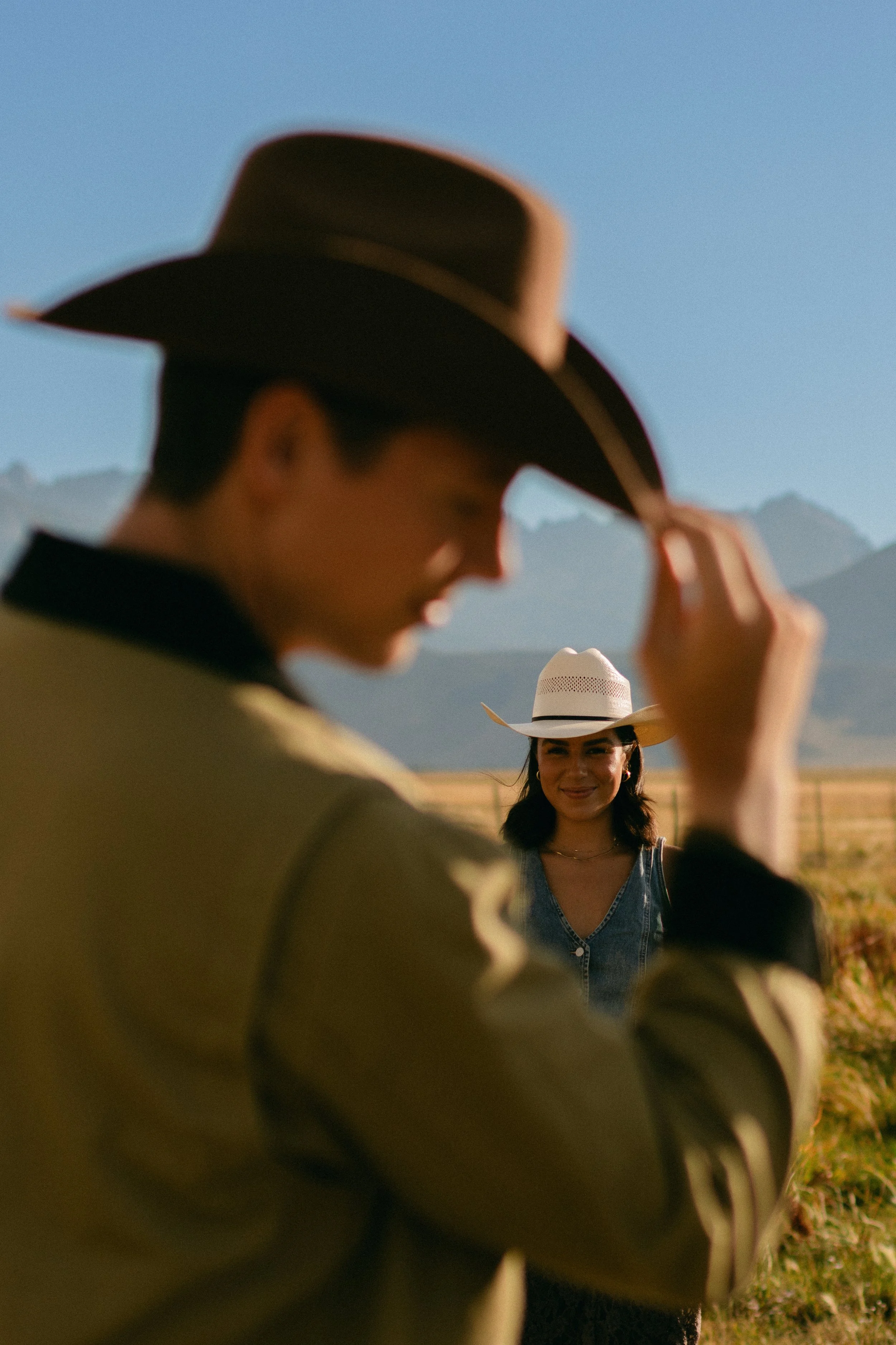 A man in a cowboy hat, slightly out of focus, holds his hat while a woman in a white wide-brimmed hat, denim shirt, and gold jewelry stands in the background smiling with mountains and a clear sky behind her.