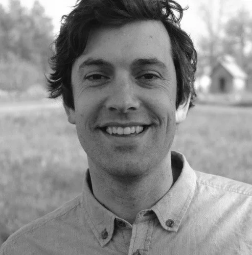 A smiling young man with messy hair in an outdoor setting with trees and a house in the background.