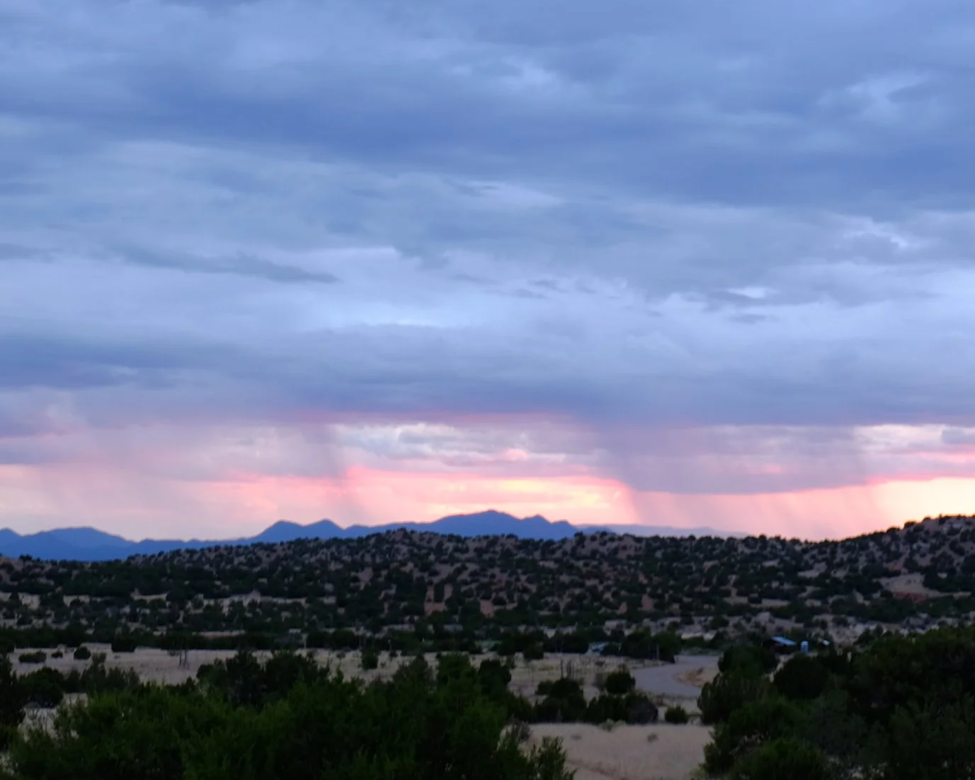 Scenic landscape with rolling hills and mountains in the distance, under a multi-colored sky with dark gray clouds and a pinkish sunset.
