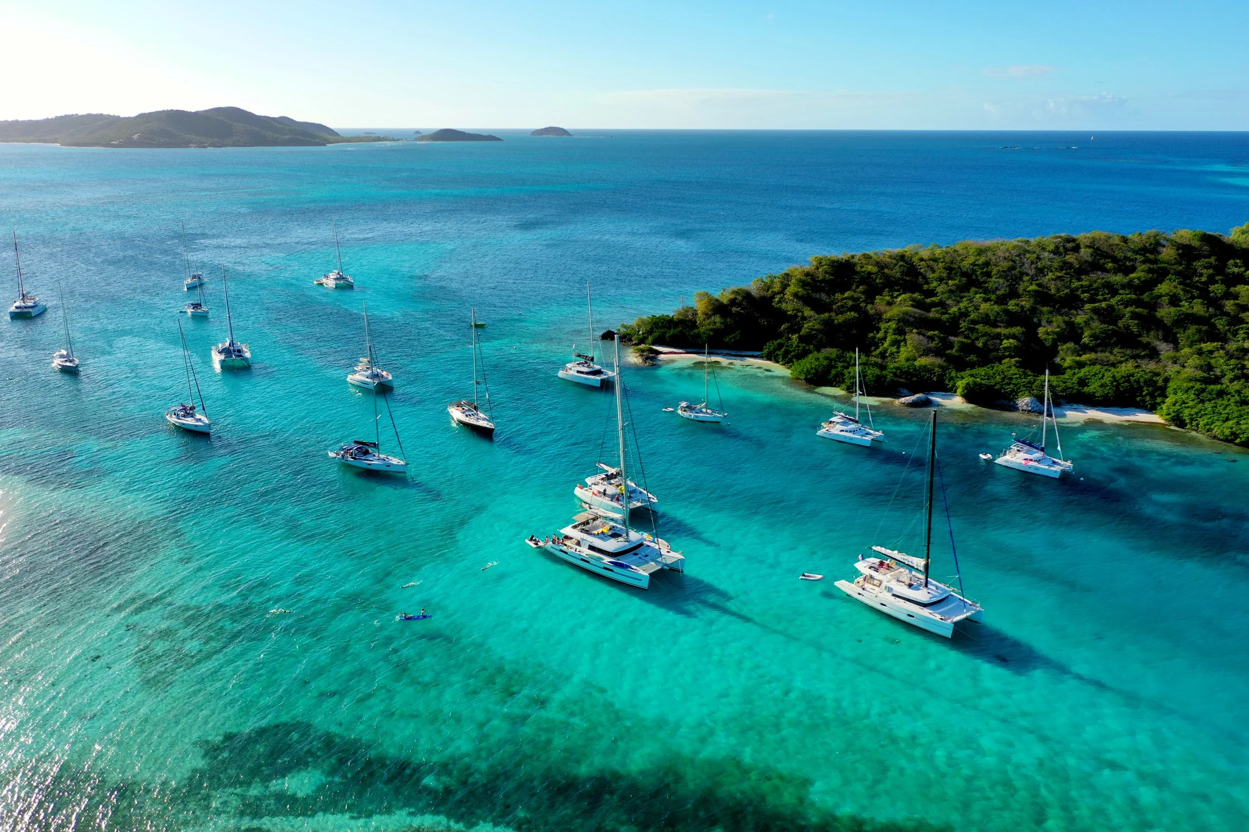 Aerial view of multiple sailboats and catamarans anchored in turquoise waters near a small island with lush greenery, under a clear blue sky.