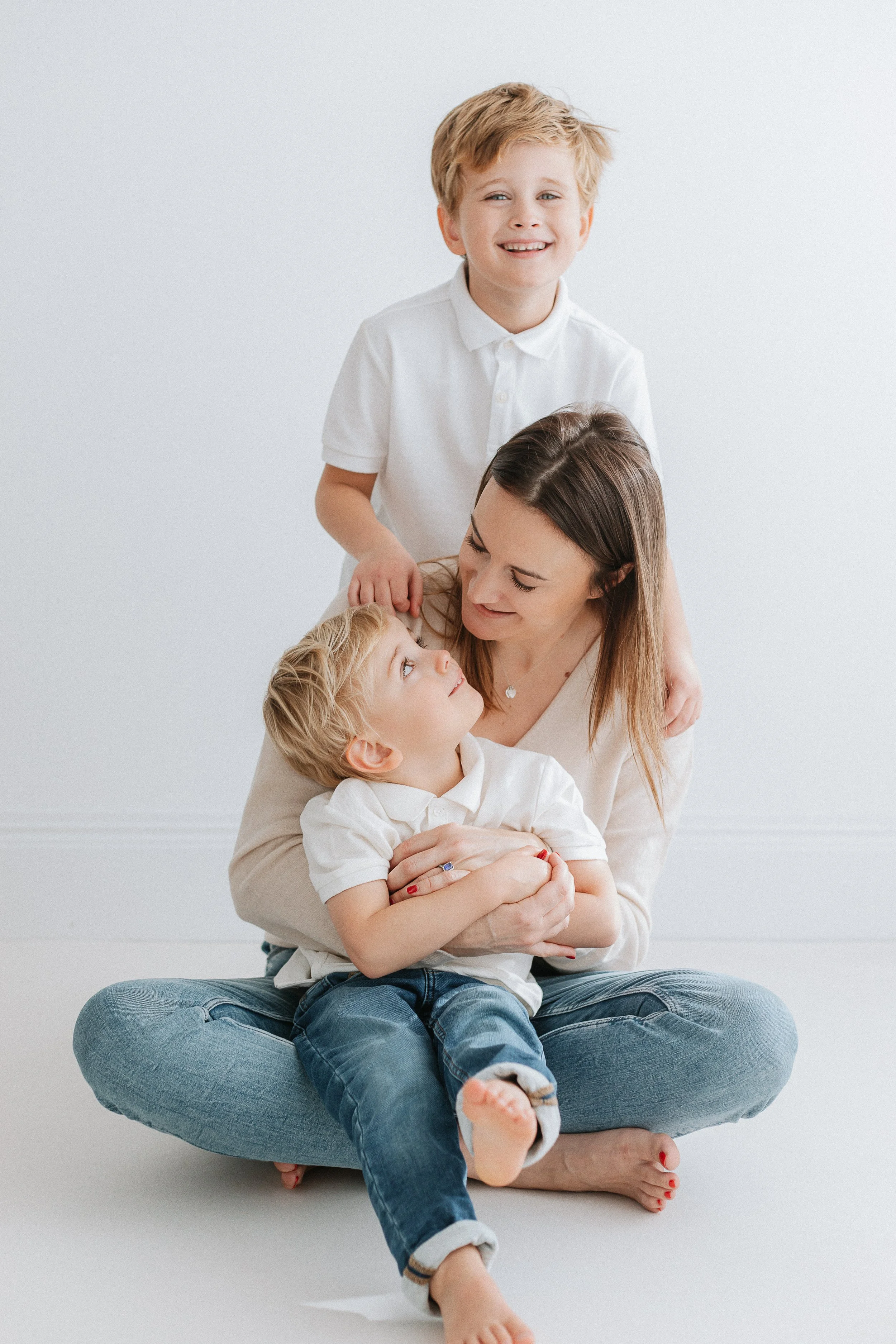 A woman sitting cross-legged on the floor, holding a boy on her lap. A smiling boy with blonde hair is standing behind them, resting his hand on the woman's head. The group is dressed in white shirts and jeans, with a plain white background.