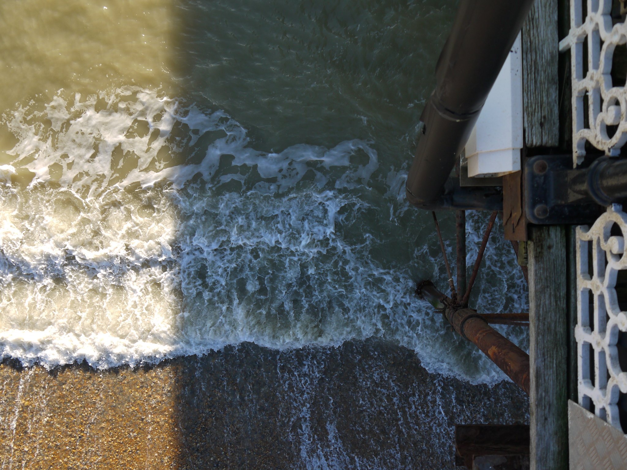 Overhead view of waves crashing on a sandy beach next to a pier structure with metal railings.