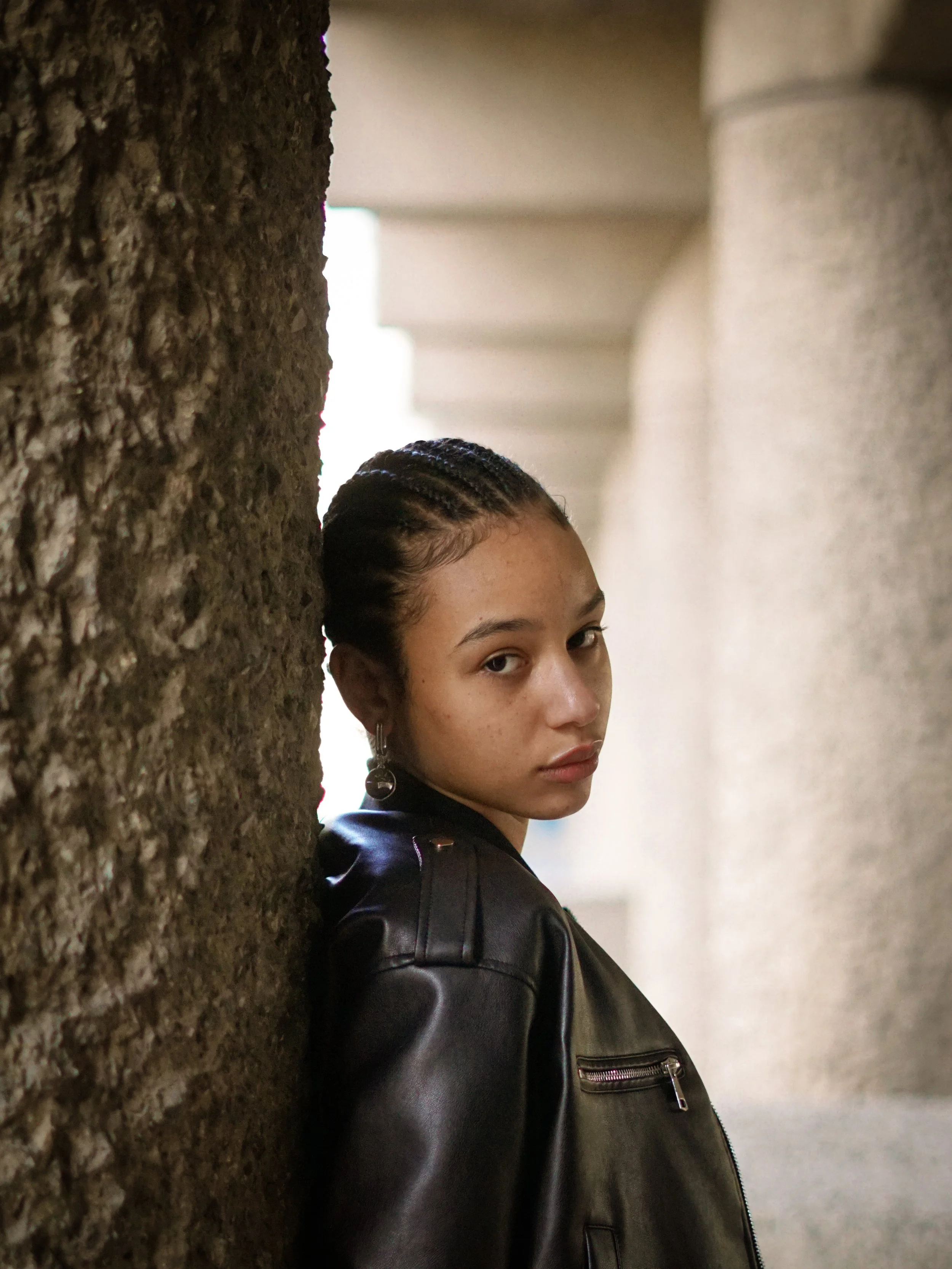 Person with braided hair wearing a black leather jacket, leaning against a textured stone wall and looking at the camera, with blurred architectural columns in the background.