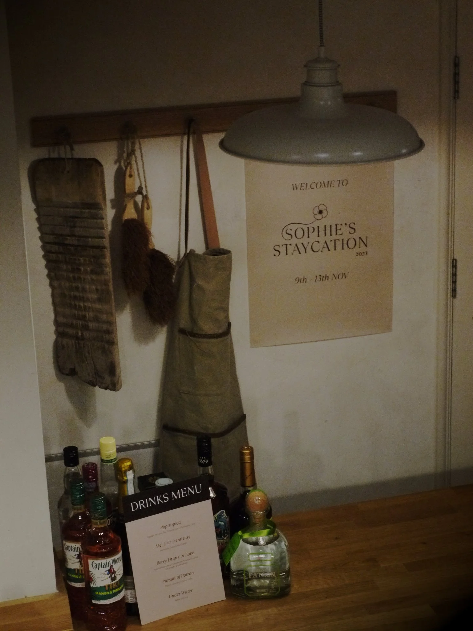 Indoor setting with a "Welcome to Sophie's Staycation" sign dated 9th to 13th November. Below the sign is a collection of liquor bottles, including brands like Captain Morgan and Patrón. A drinks menu is visible with cocktail names. Hanging on the wa