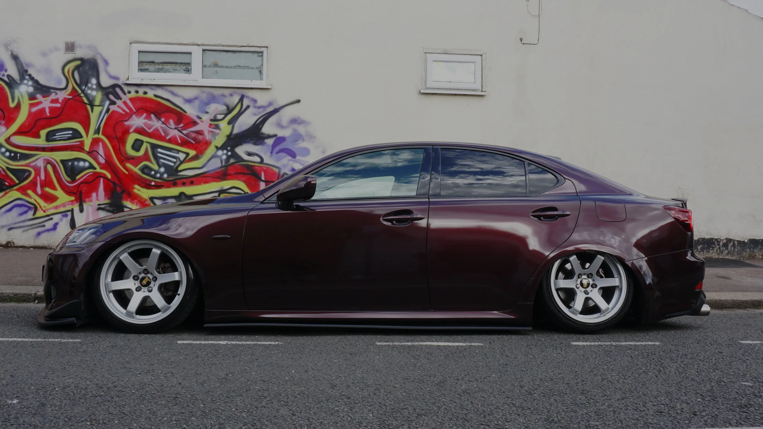 Slammed dark-colored sedan with custom wheels parked in front of a graffiti-covered wall.
