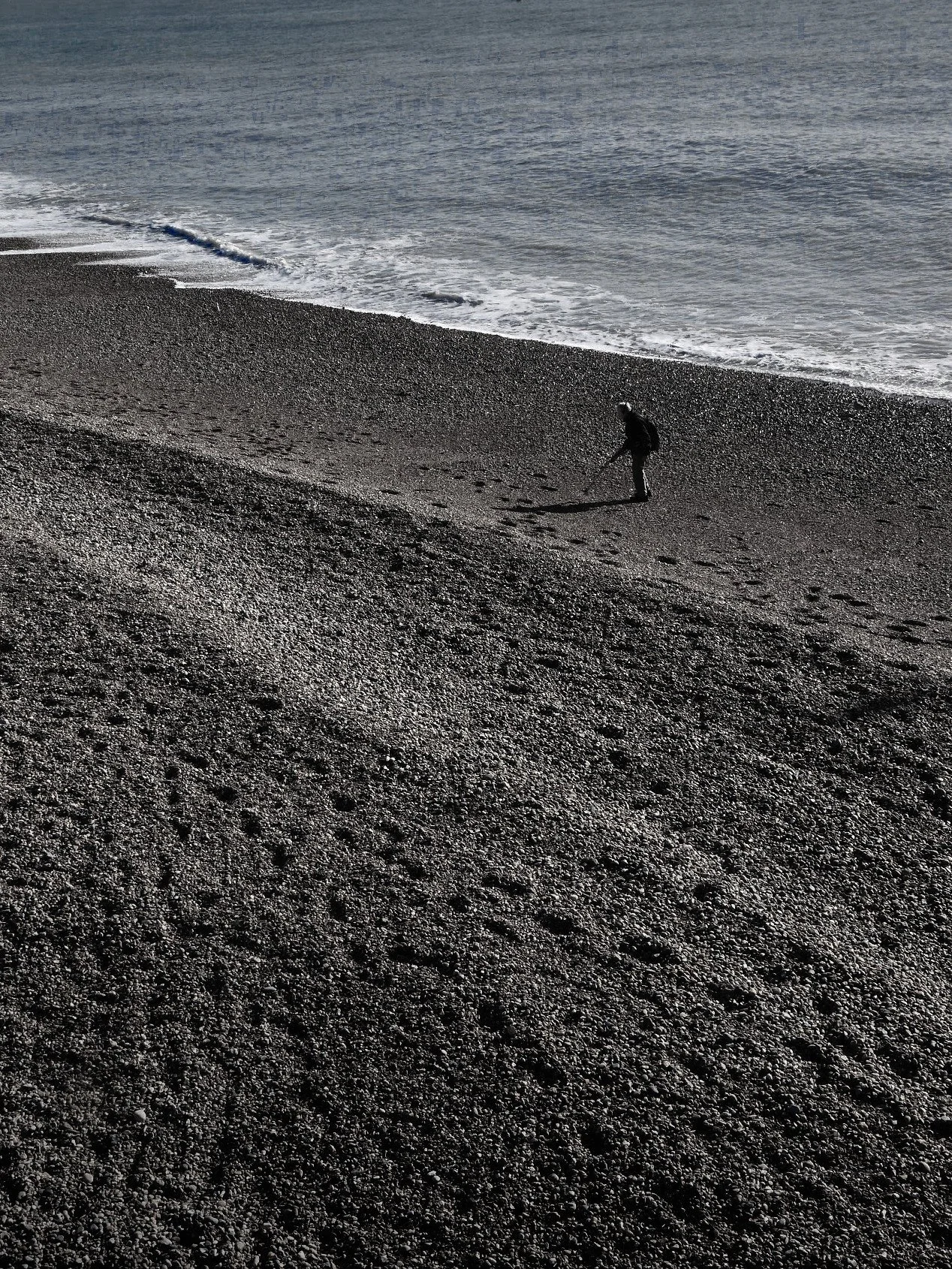 Person with a metal detector walking on a rocky beach near the shoreline.