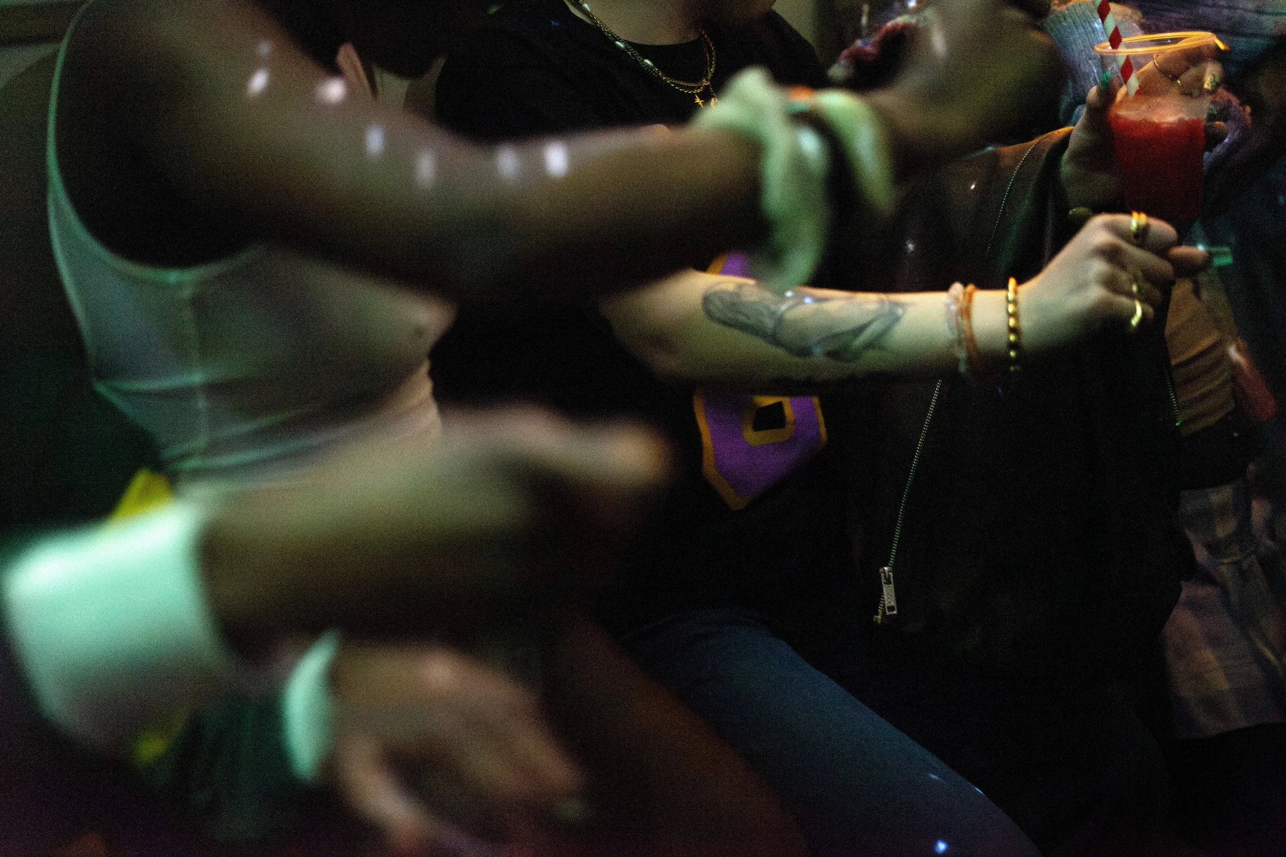 Close-up of people dancing in a dimly lit setting, featuring arms raised, colorful bracelets, a visible tattoo, and a hand holding a red drink with a straw.