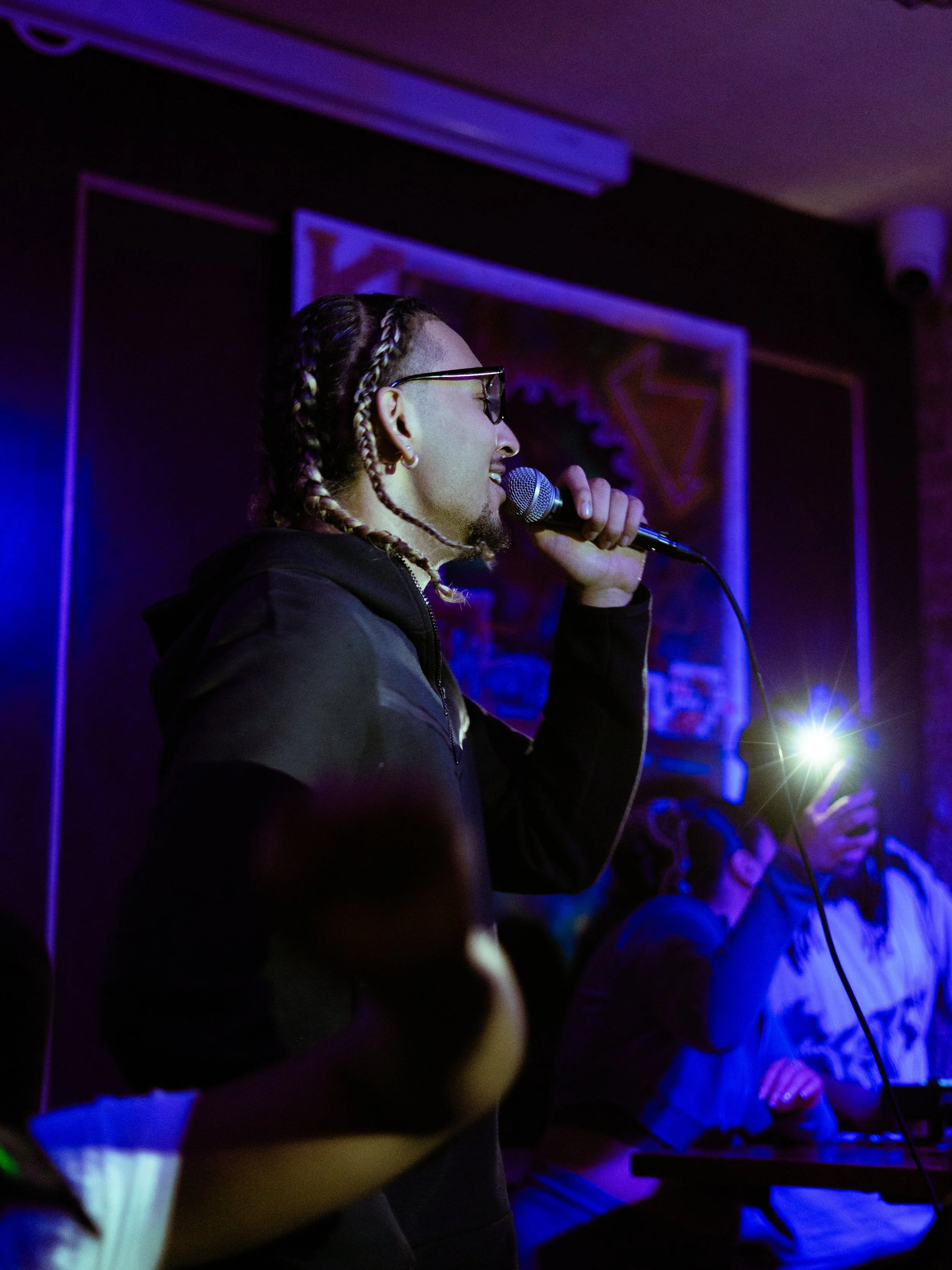 Person with braids performing on stage with a microphone, audience taking pictures.