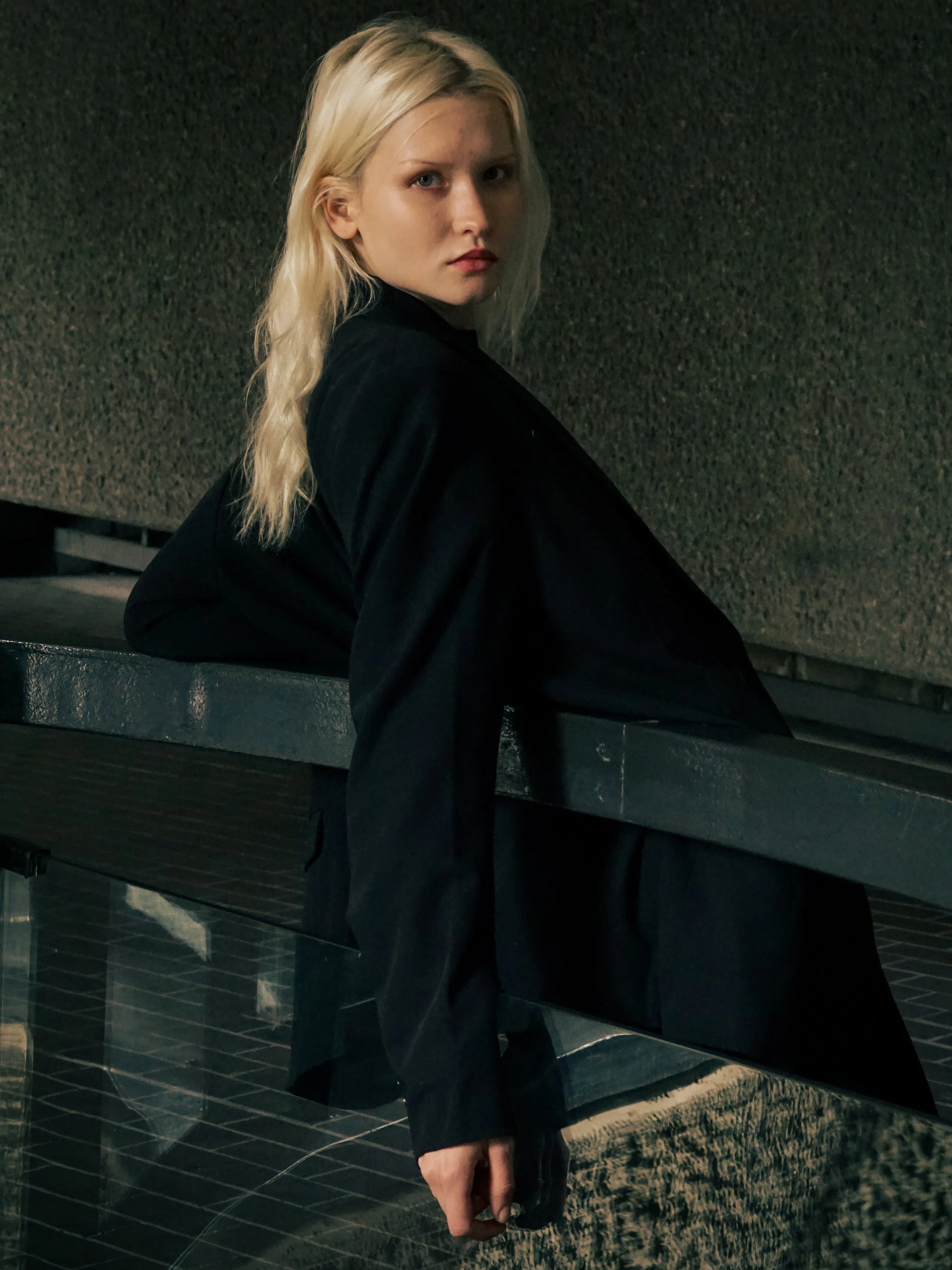 Person with long blonde hair leaning on railing, wearing black jacket, in dimly lit setting.