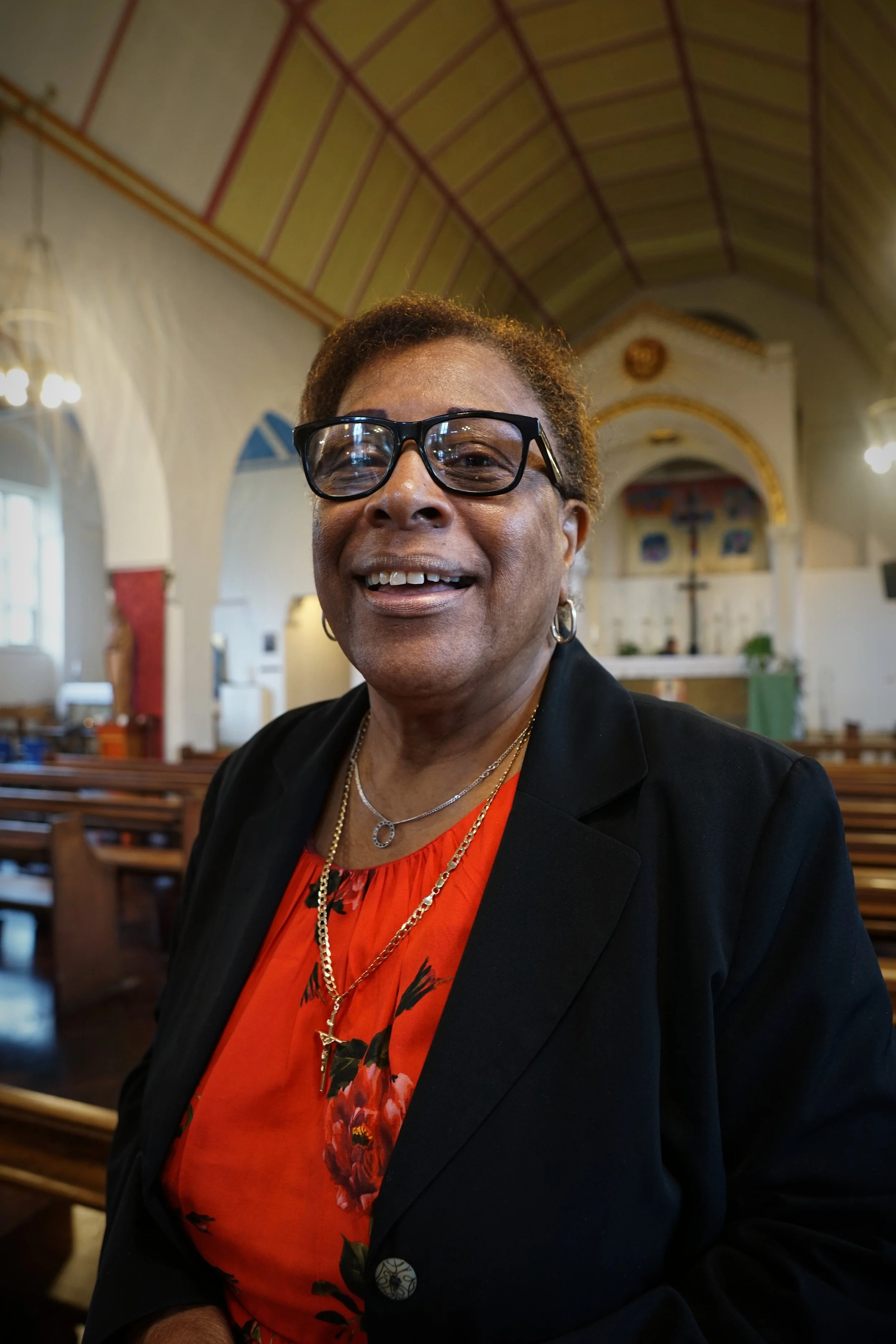 Person smiling in a church, wearing glasses and a red floral shirt with a black blazer.