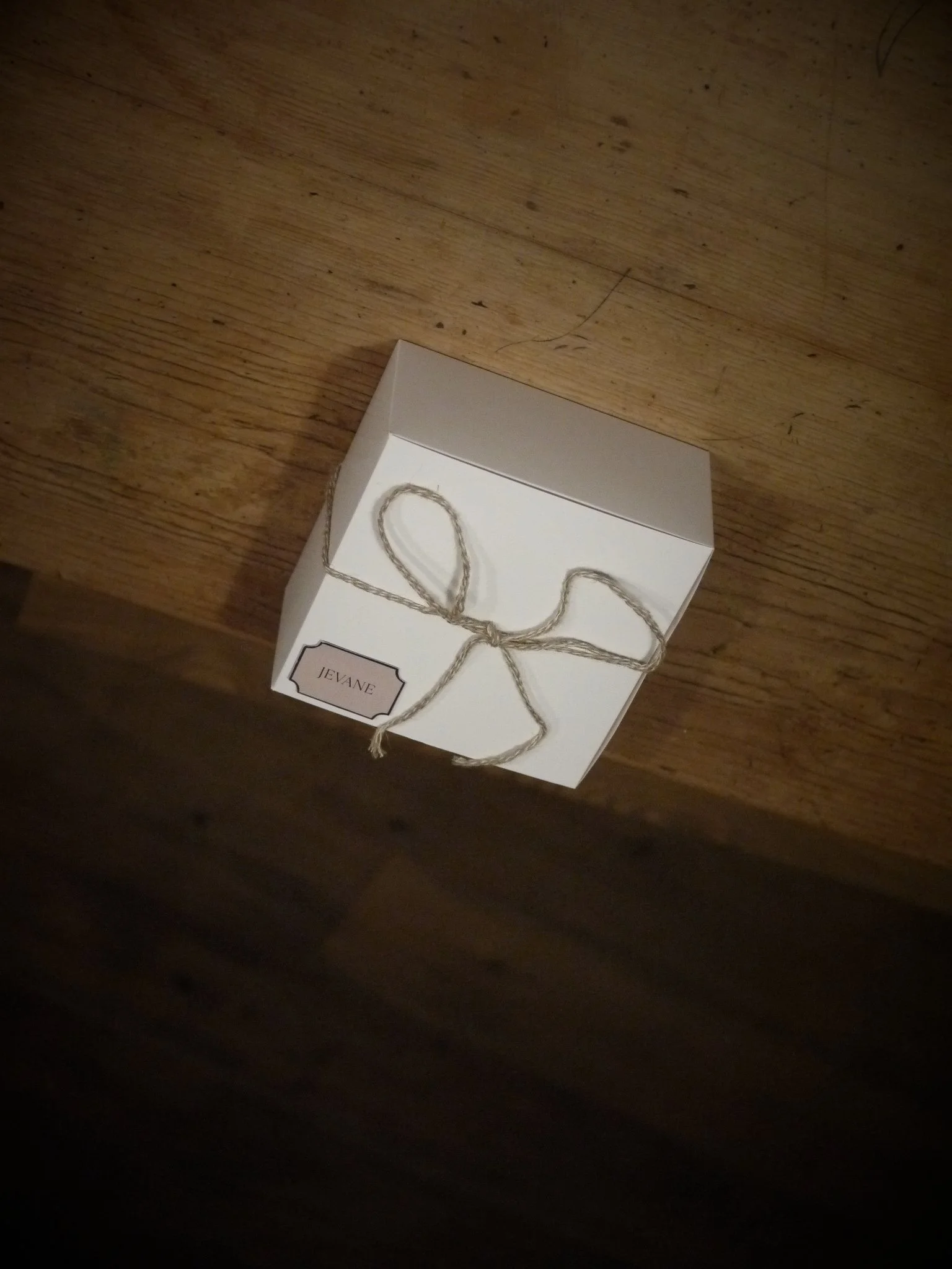 A small white gift box tied with twine, labeled "Jevane," resting on a wooden surface.