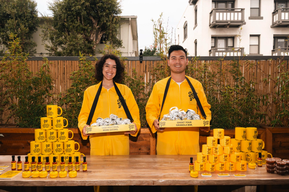 Two people dressed in yellow jackets standing behind a table filled with yellow mugs, small bottles, and jars, outdoors in front of a wooden fence and green plants, smiling at the camera.