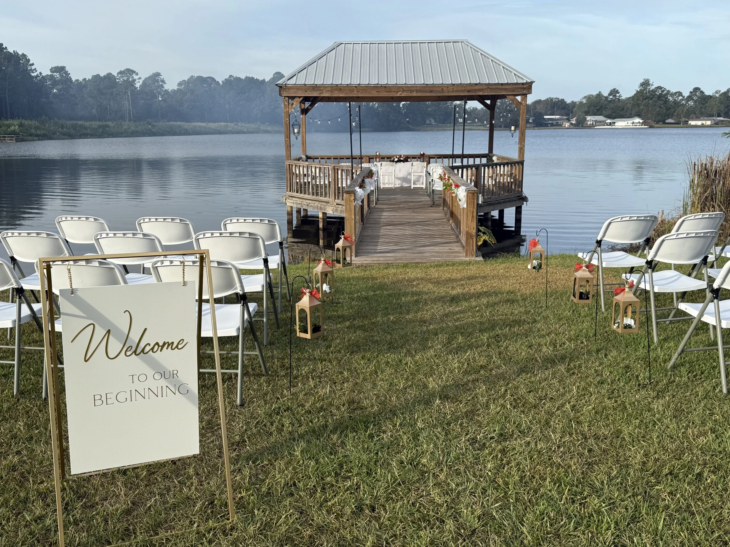 A lakeside wedding setup with white chairs, a wooden dock with swings, and a welcome sign on a grassy area.