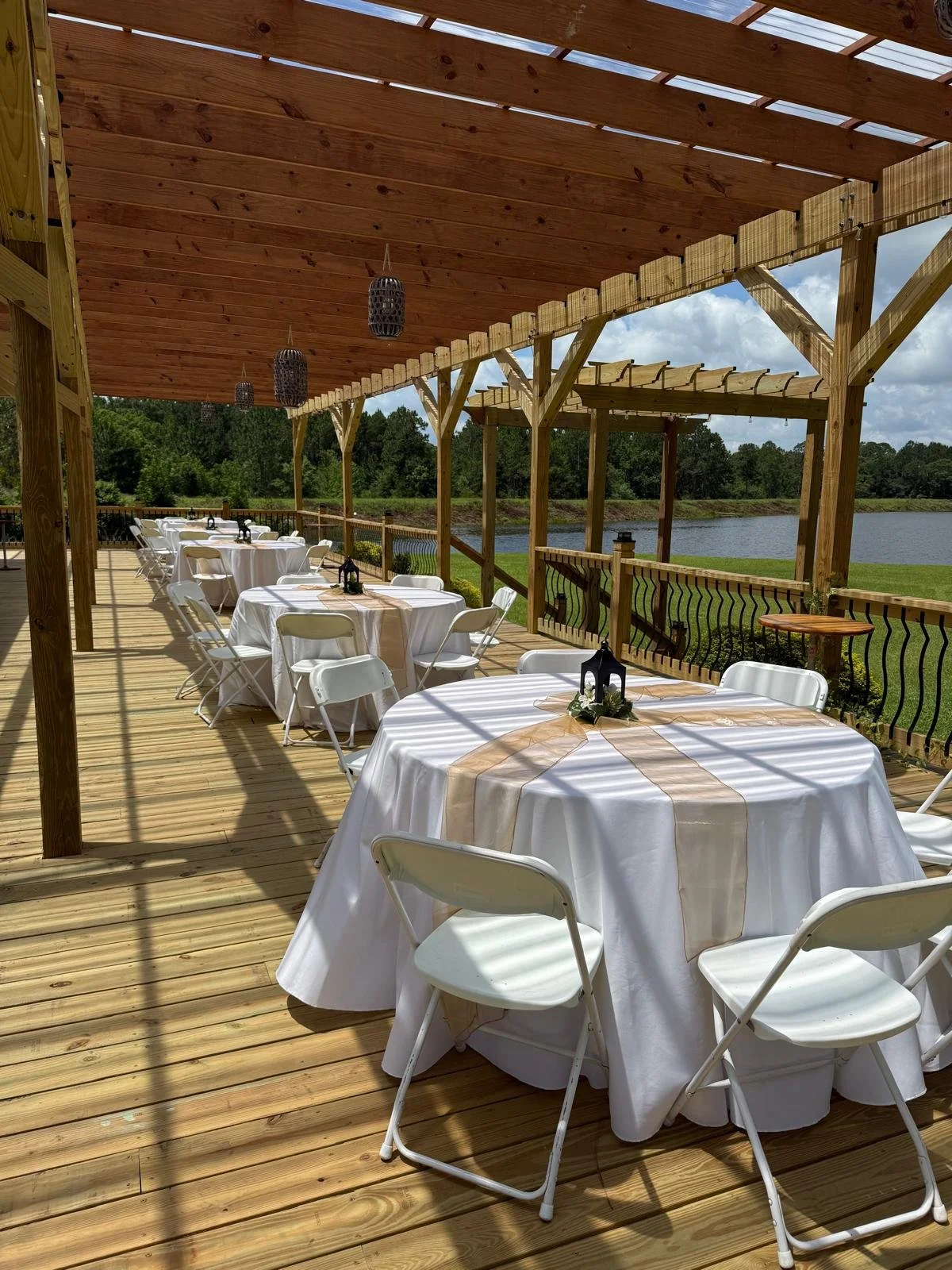Outdoor wooden deck with round tables covered in white tablecloths, decorated for an event, each with a beige runner and a small lantern centerpiece. White folding chairs surround the tables. The deck has a wooden railing overlooking a scenic view of