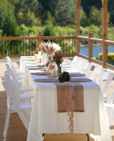 Outdoor dining table set with white chairs, candles, and floral centerpiece on a wooden deck, overlooking trees and water in the background.