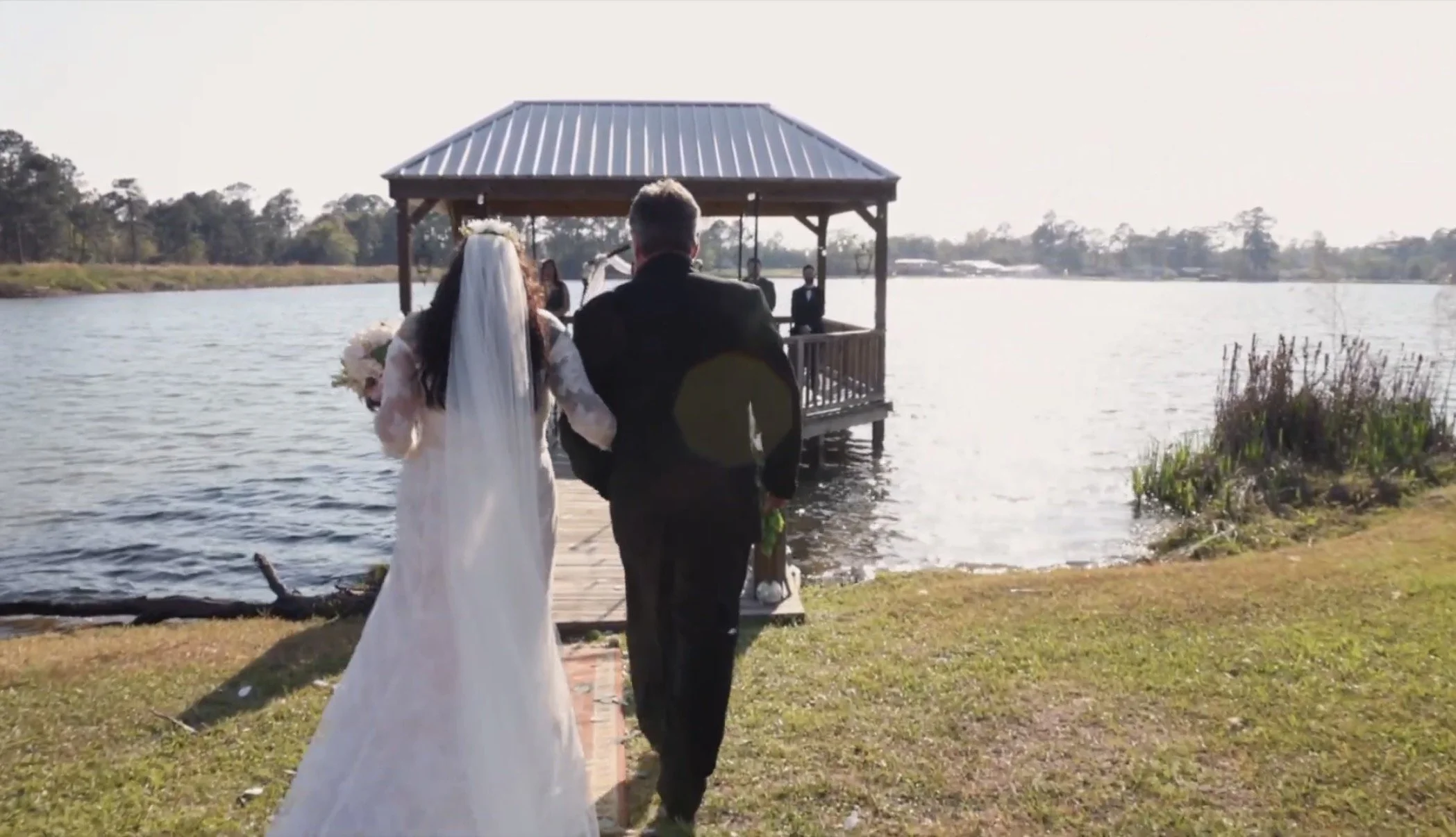 A bride and groom walking on a dock towards a gazebo over a lake, with a priest or officiant standing inside the gazebo.
