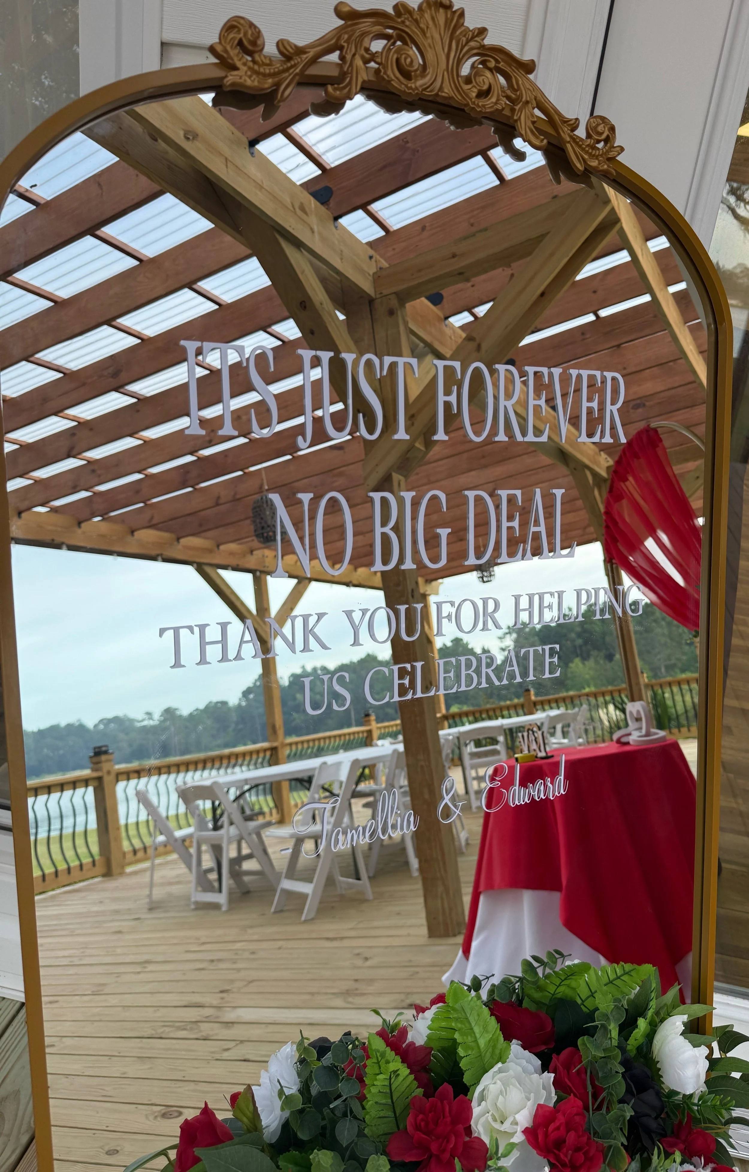 A wedding or celebration sign on a mirror with outdoor deck and wooden gazebo in the background. The mirror has decorative gold trim and an inscription reading "It is just forever no big deal thank you for helping us celebrate." There are white chair