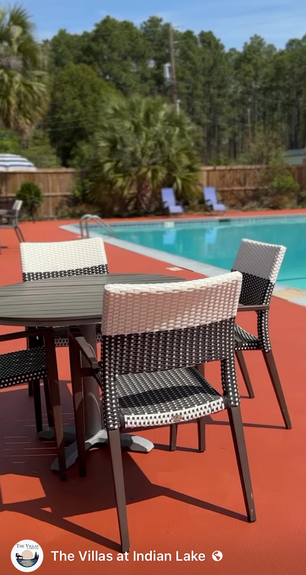 View of a backyard with a swimming pool, a patio with wicker chairs and a table, surrounded by a wooden fence, with trees in the background and a partly cloudy sky.