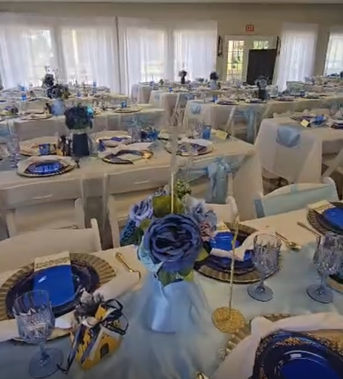 Elegant banquet setup with white tablecloths, navy and gold dinnerware, floral centerpieces with roses, and gold accents in a spacious room with large windows and white curtains.