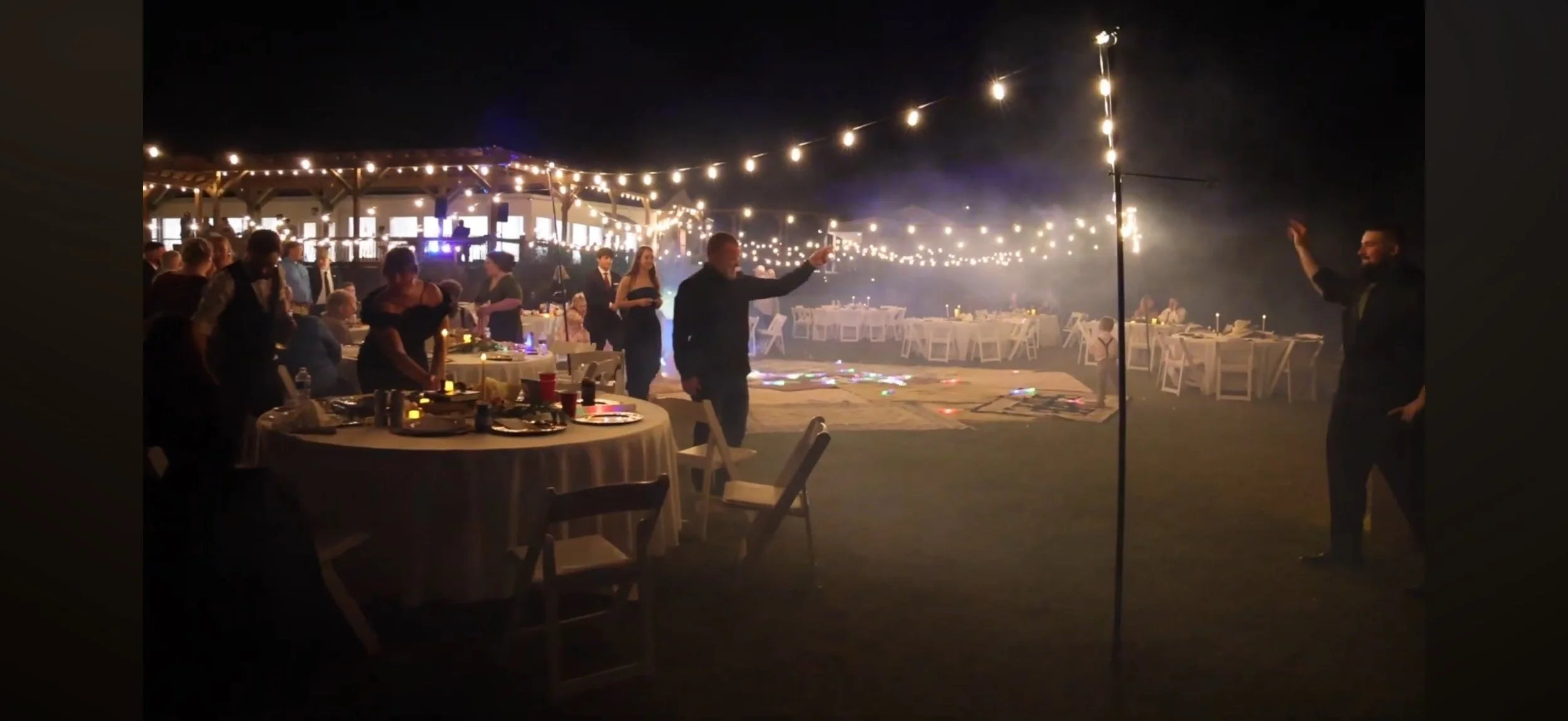 Nighttime outdoor party with string lights, decorated tables, and people dancing or socializing, with one person on a dance floor and another holding a microphone, in a festive setting.