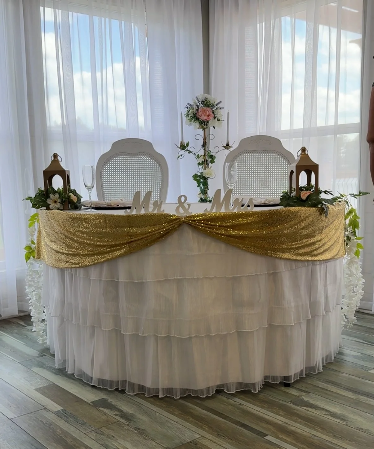 Wedding reception table decorated with gold and white fabric, floral arrangements, lanterns, and 'Mr. & Mrs.' sign, set against sheer white curtains and wooden floor.