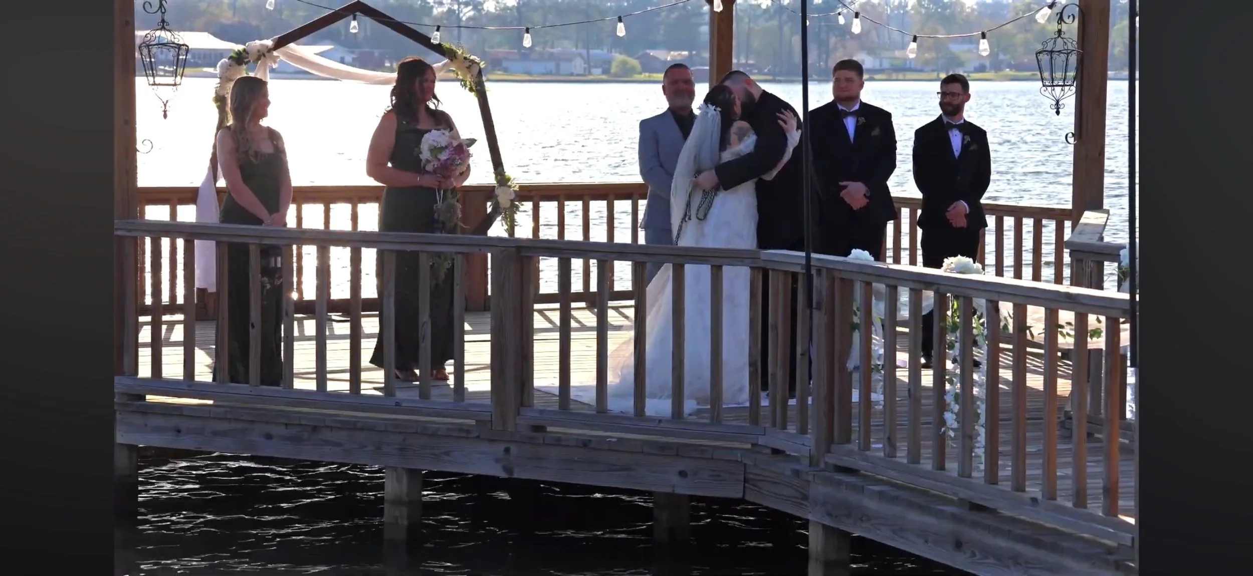 A wedding ceremony taking place on a wooden dock by a lake, with a bride and groom kissing, surrounded by their wedding party and officiant, with string lights overhead and scenic water in the background.