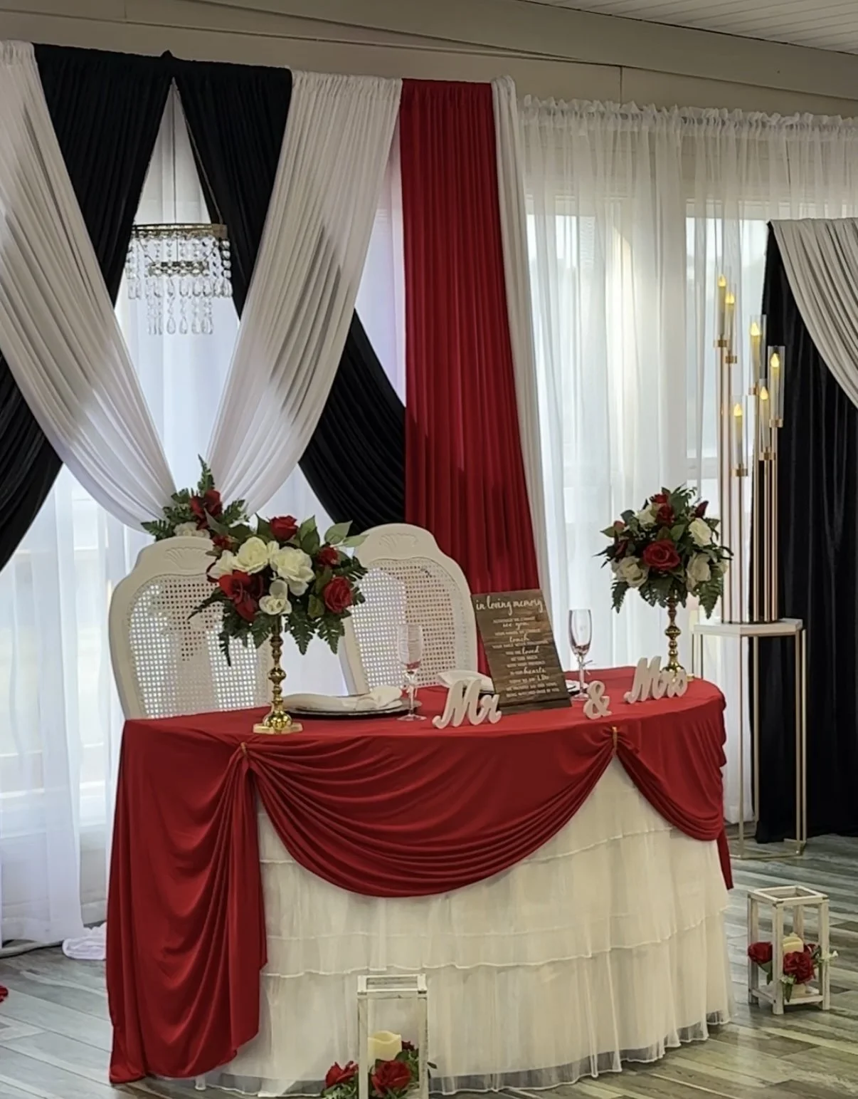 Wedding reception table decorated with red and white drapery, floral arrangements of red and white roses, and signs reading 'Mr' and 'Mrs.' with a festive and romantic setup.