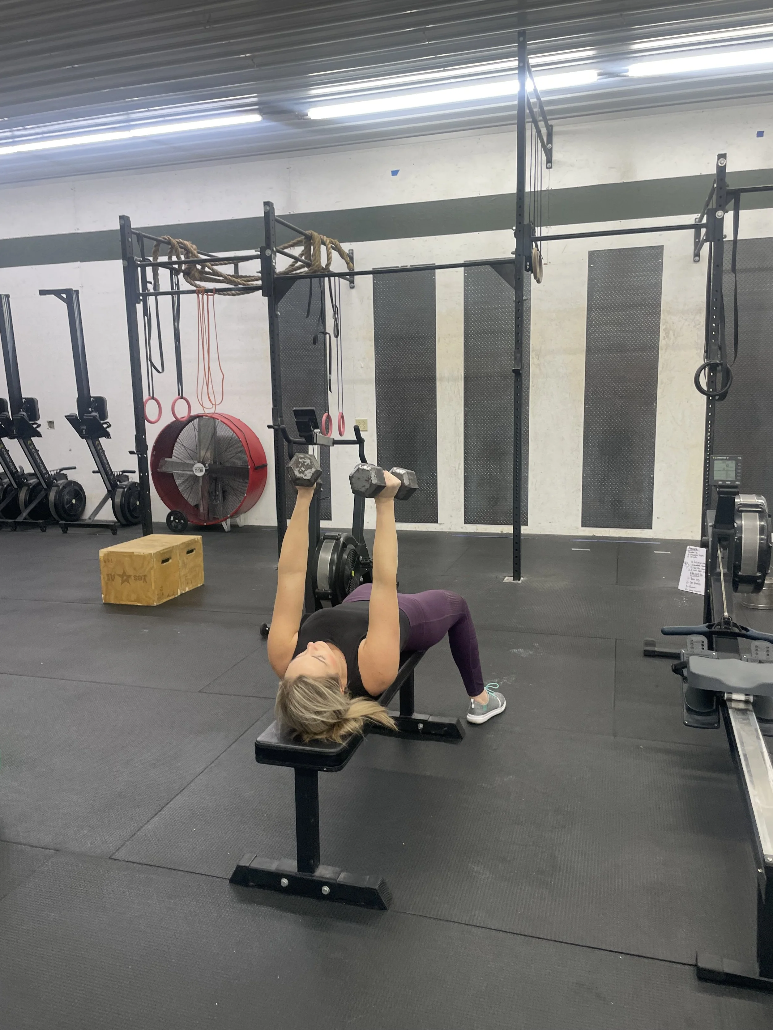 Woman lying on a bench performing a dumbbell chest press exercise in a gym. She is holding a dumbbell in each hand with her arms extended upward. The gym has various equipment including rowing machines, a large fan, gymnastic rings, ropes, and a plyometric box.