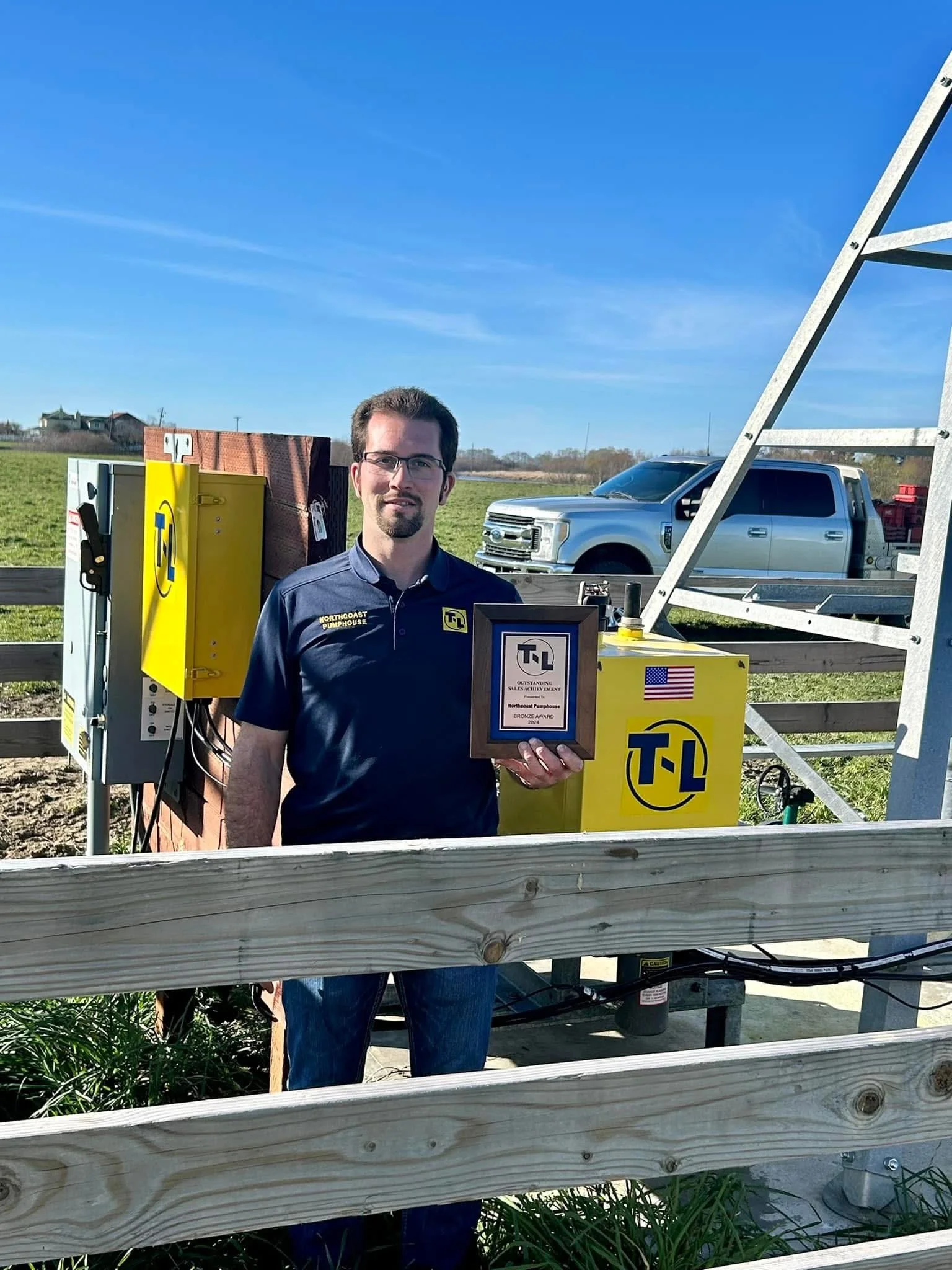 A man holding an award plaque standing outdoors near yellow utility equipment and a silver pickup truck, with a blue sky and green field in background.