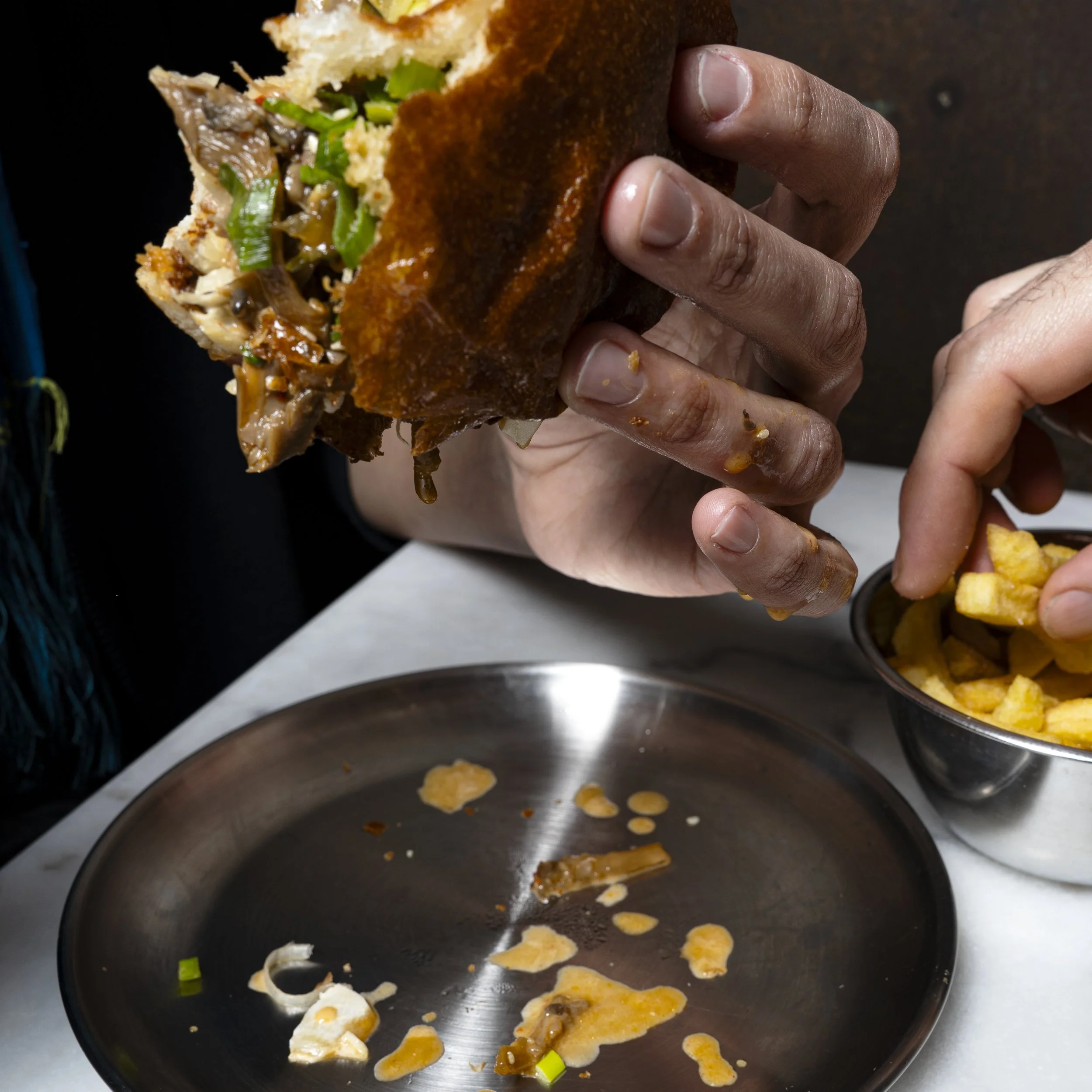Hands holding a vegan burger with fresh green vegetables and soft buns, served with a small bowl of fries on a table.