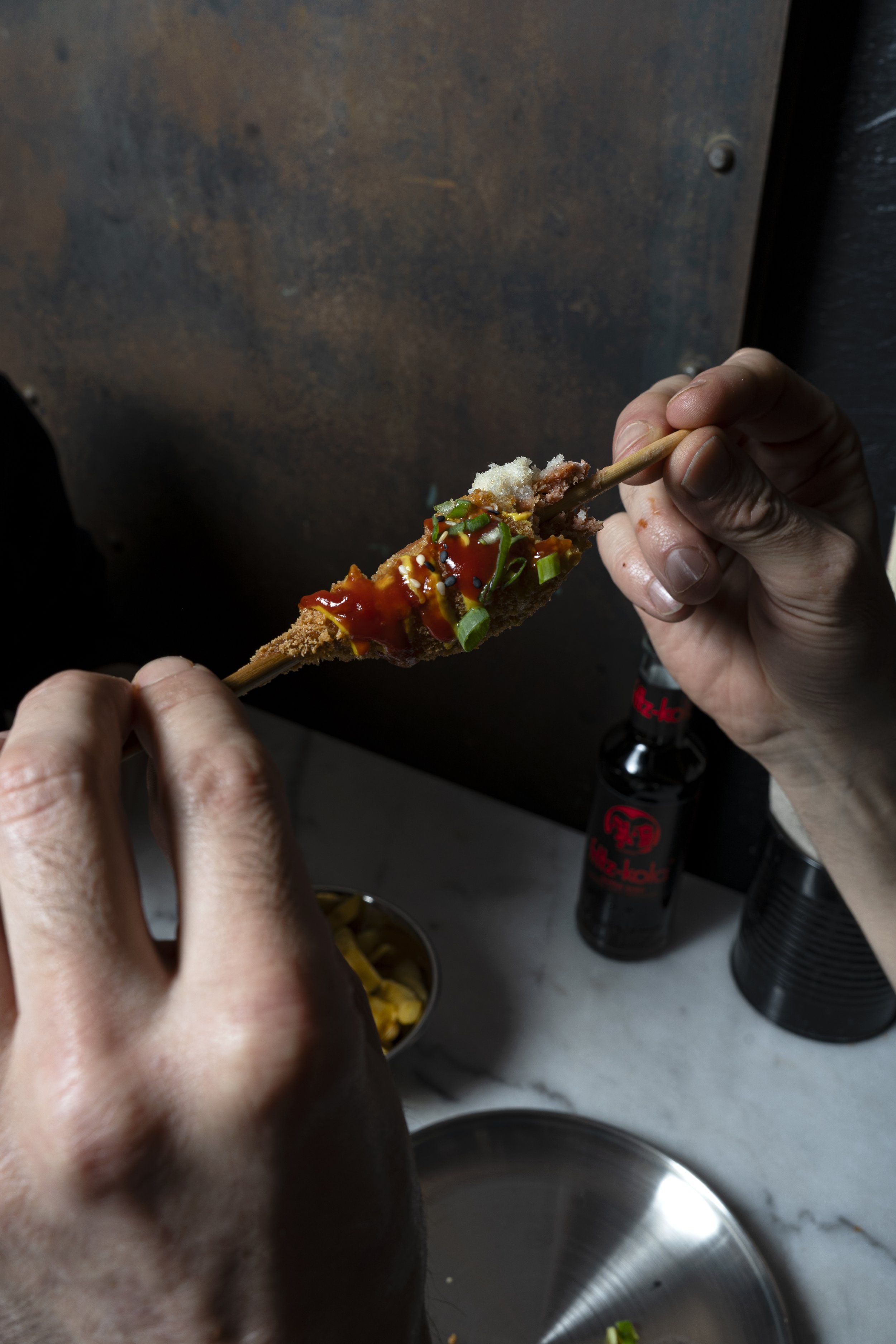 A hand holding a vegan skewer coated in sauce with plant-based sausage, vegetables, and spices in a kitchen setting.