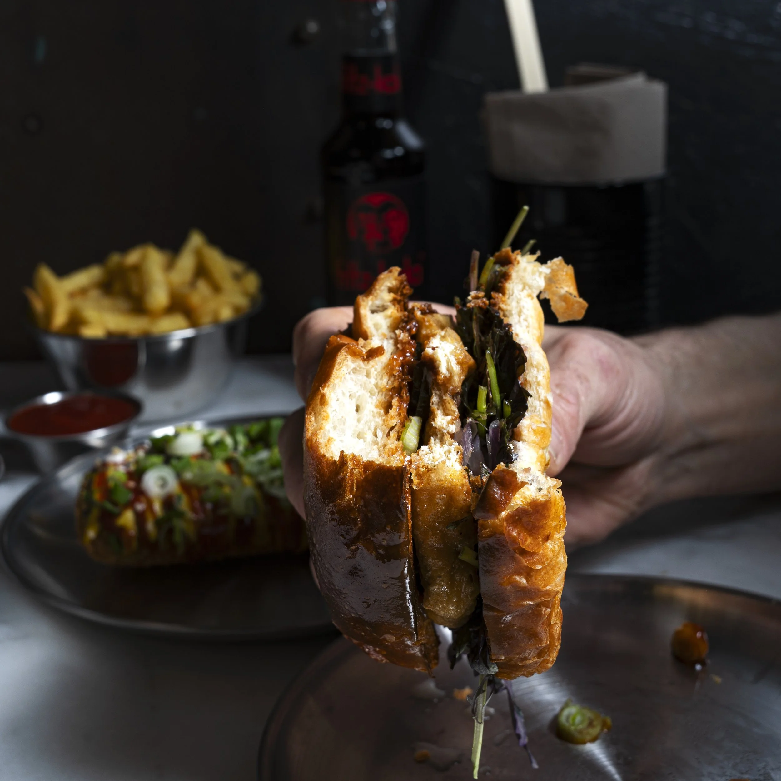 A hand holding a vegan burger with plant-based meat, fresh green vegetables, and sauce. In the background, a bowl of fries with ketchup, vegan corn dog on a plate, and a bottle of soda on the table.