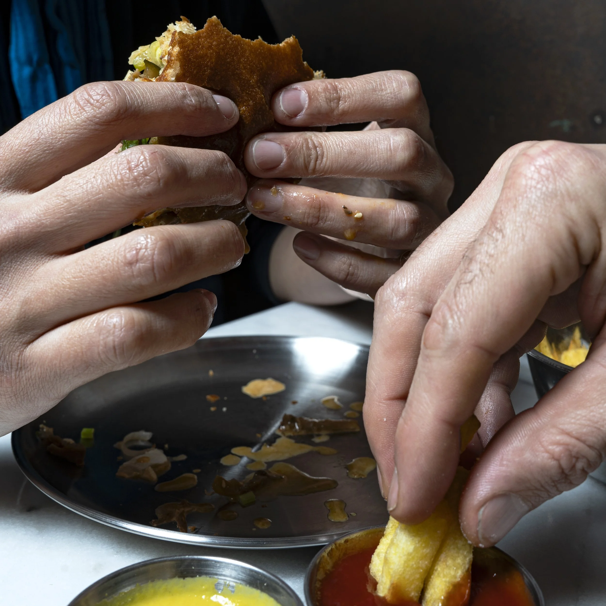 Two hands holding a vegan burger made with homemade bread, falafel, and fresh vegetables, above a metal plate with food remnants and small bowls of ketchup and sweet mustard sauces.