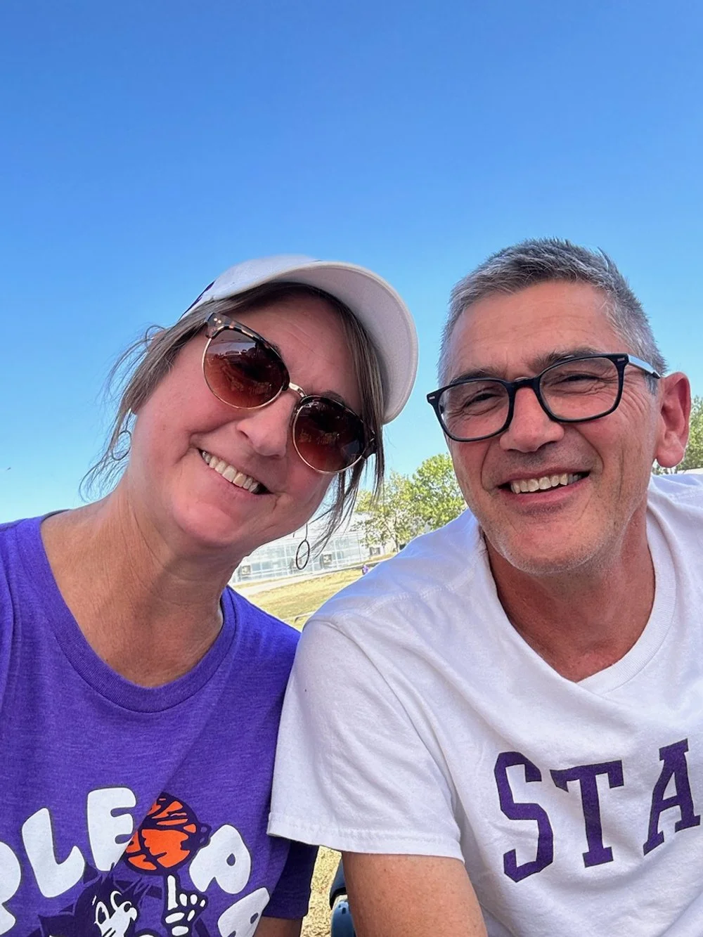 A smiling woman with sunglasses and a white cap, and a smiling man with glasses, sitting outdoors on a sunny day with a clear blue sky and green trees in the background.