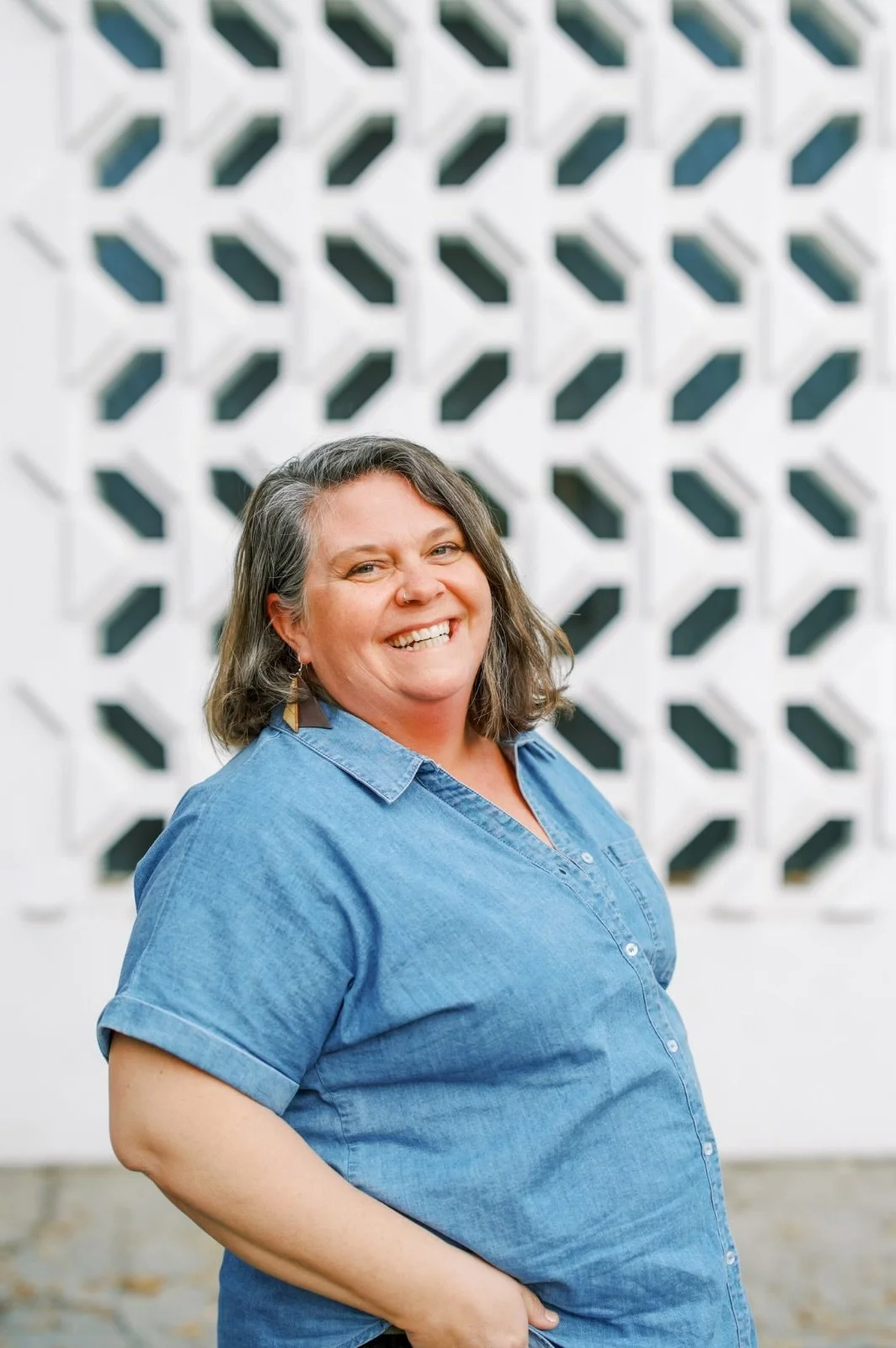 A smiling woman with shoulder-length gray hair wearing a denim shirt standing outdoors, with a geometric patterned white wall in the background.