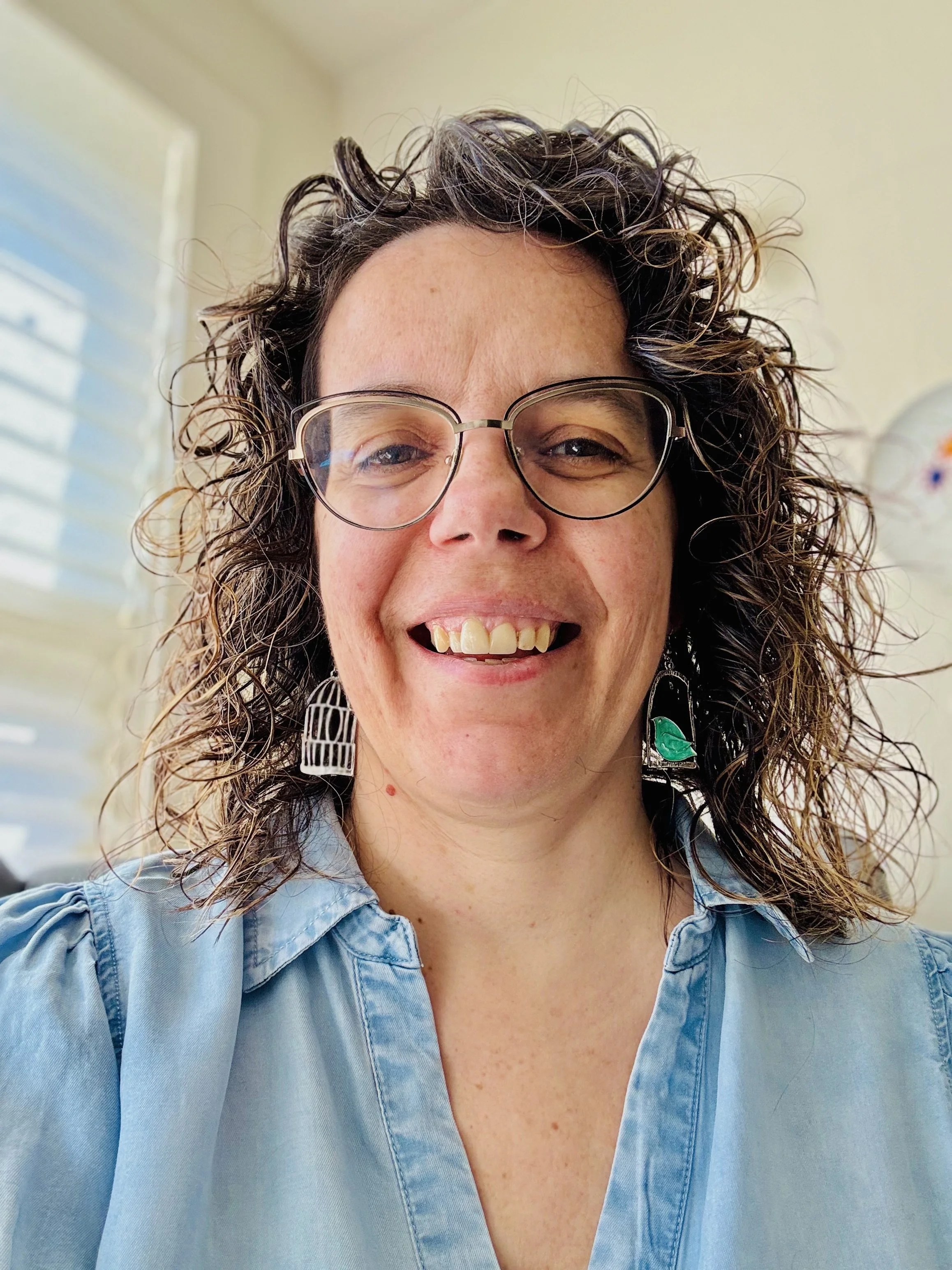 Smiling person with curly hair, wearing glasses and earrings, dressed in a light blue shirt, in a well-lit room.