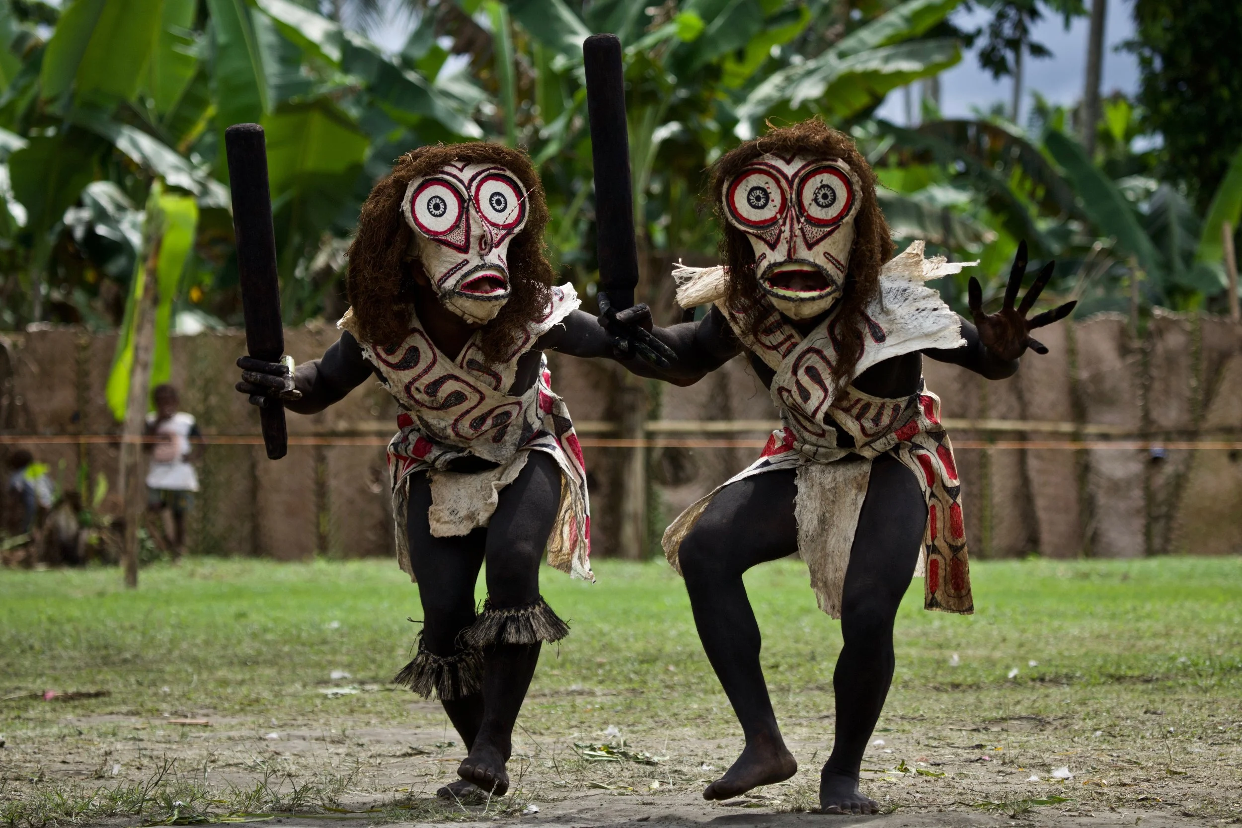 Two performers in traditional Rabaul masks and ceremonial attire dancing with wooden props during a cultural event in Papua New Guinea, set against a tropical village backdrop.