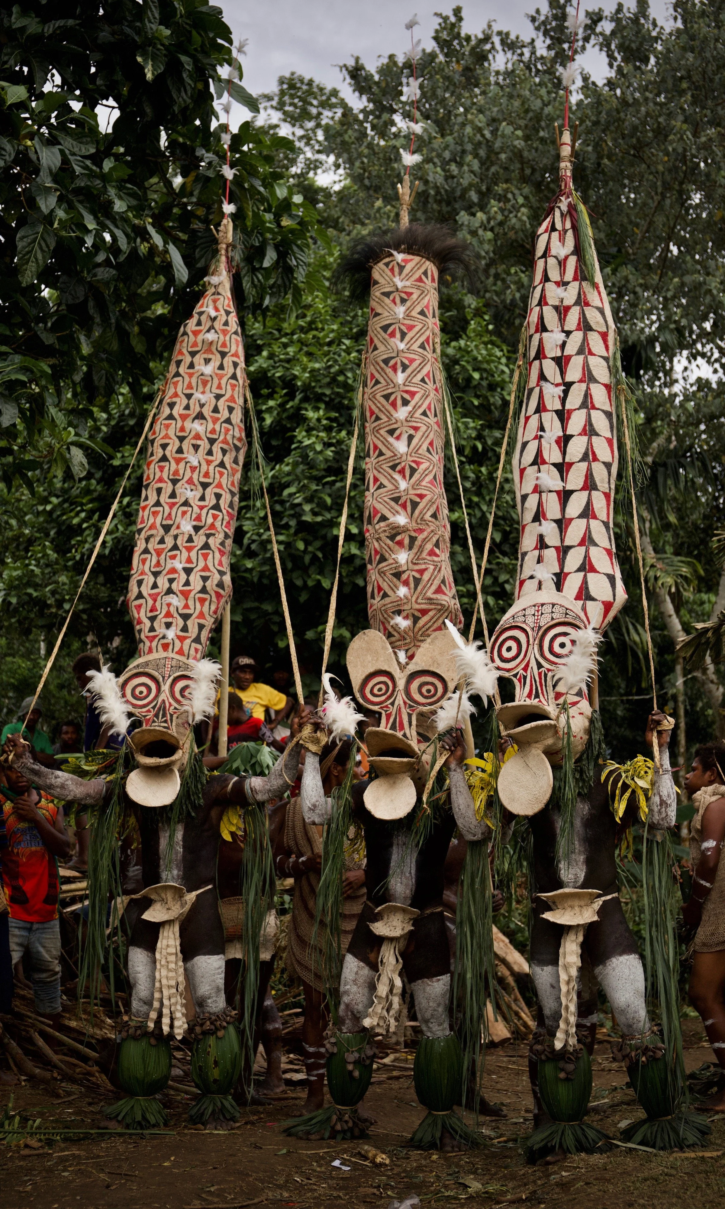 Traditional Rabaul dancers wearing towering, hand-painted masks with bold red, white, and black patterns during a cultural performance in Papua New Guinea, surrounded by onlookers in a lush village setting.
