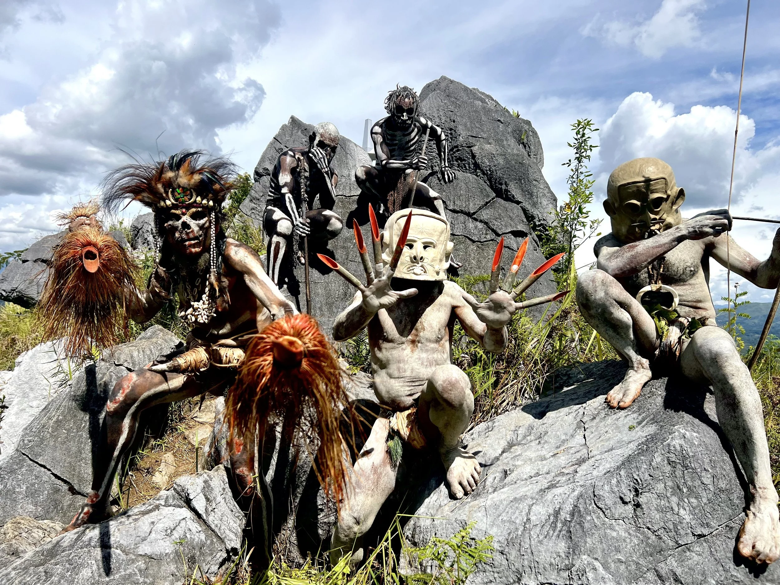 Asaro Mud Men of Papua New Guinea wearing traditional clay masks and body paint, posed with dramatic gestures and props among rocky terrain during a cultural performance.