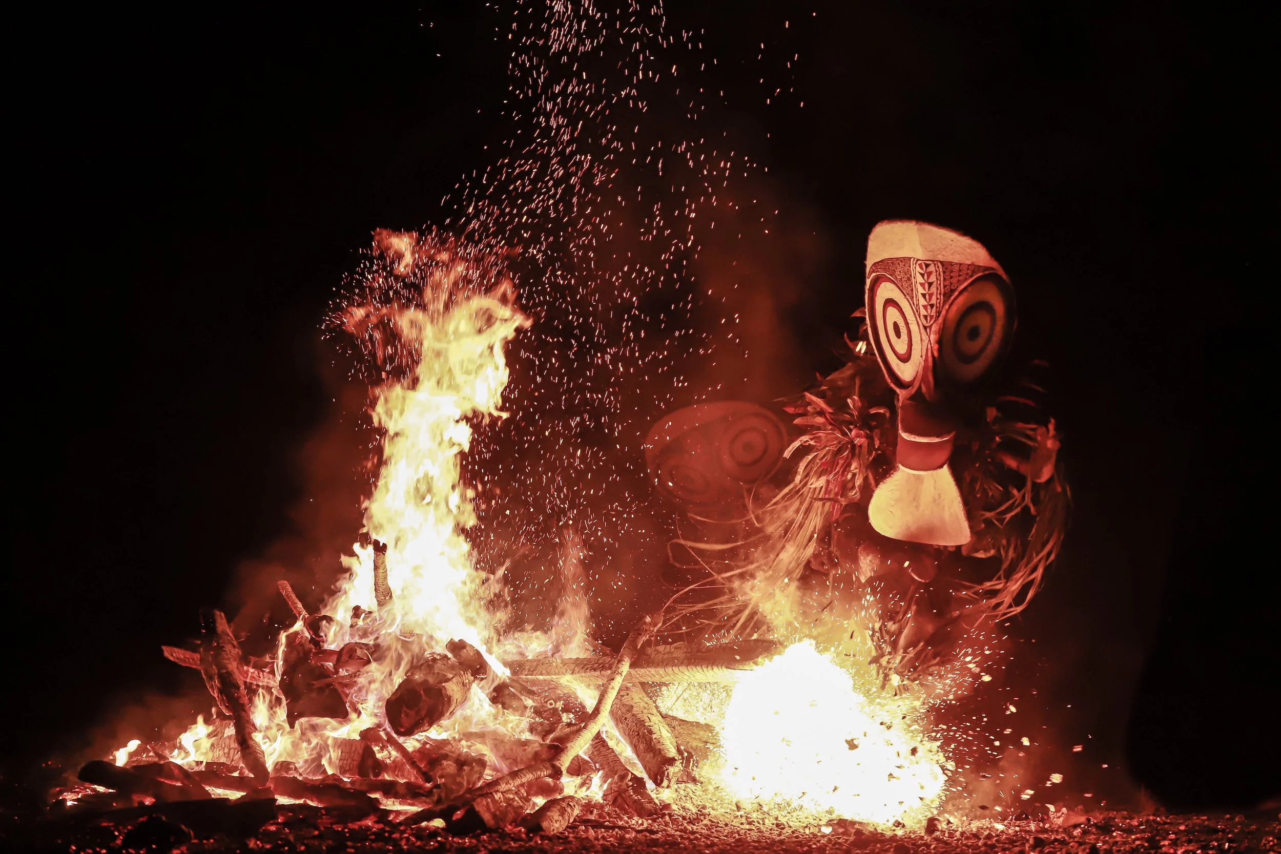 Baining fire dancer in a traditional mask and costume performing beside roaring flames at night in Papua New Guinea, with sparks and embers lighting the scene in a dramatic cultural display.