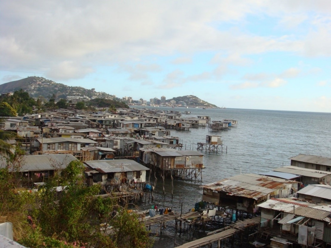 Coastal stilt houses lining the waterfront near Port Moresby, Papua New Guinea, with urban hills and the city skyline visible in the distance under a partly cloudy sky.