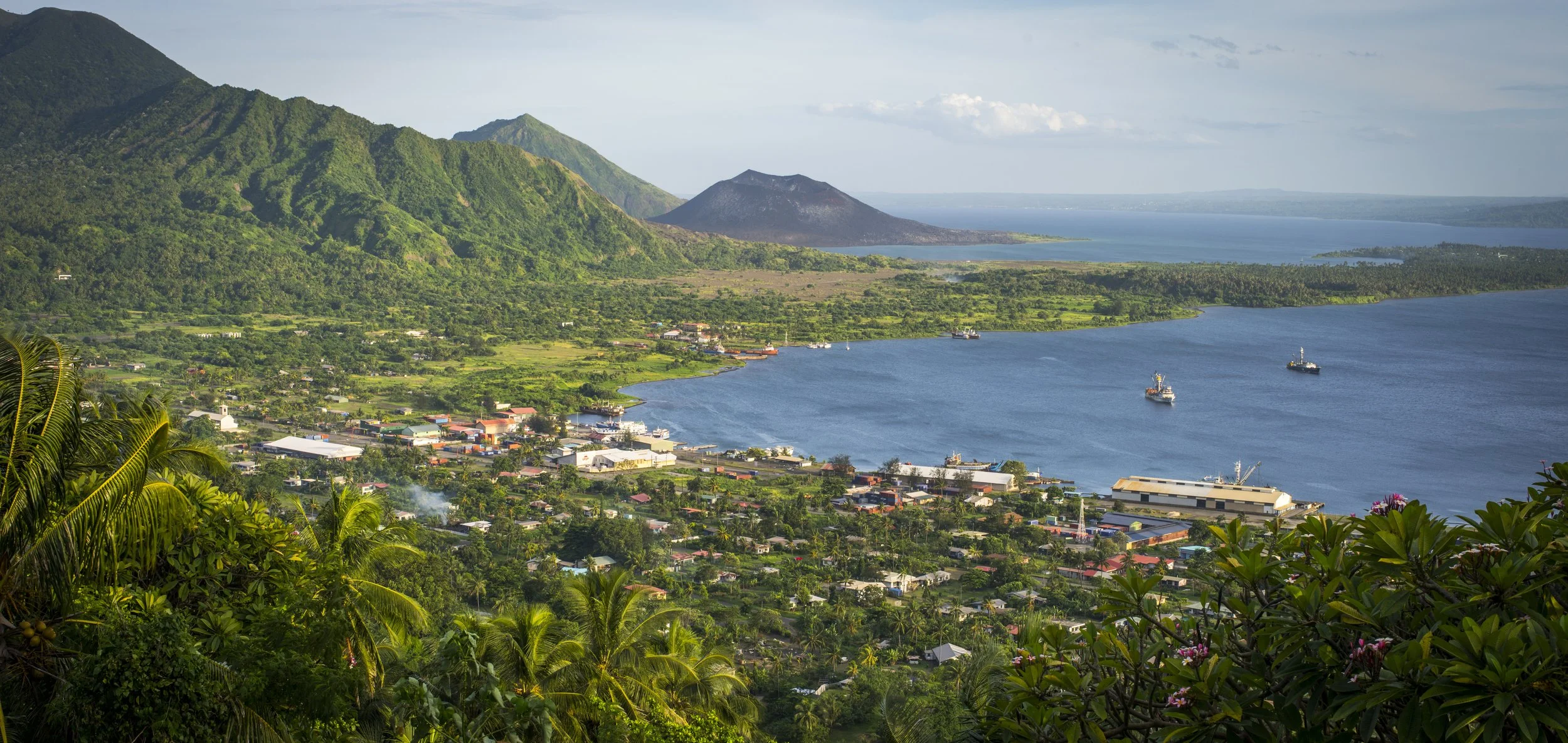 Scenic view over Rabaul town in Papua New Guinea, with tropical greenery, a calm bay, and volcanic peaks rising in the distance beneath a clear sky.