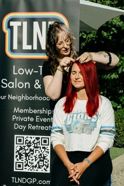 Stylist styling vivid red hair for a seated client in front of a TLND low tox salon banner in Grosse Pointe