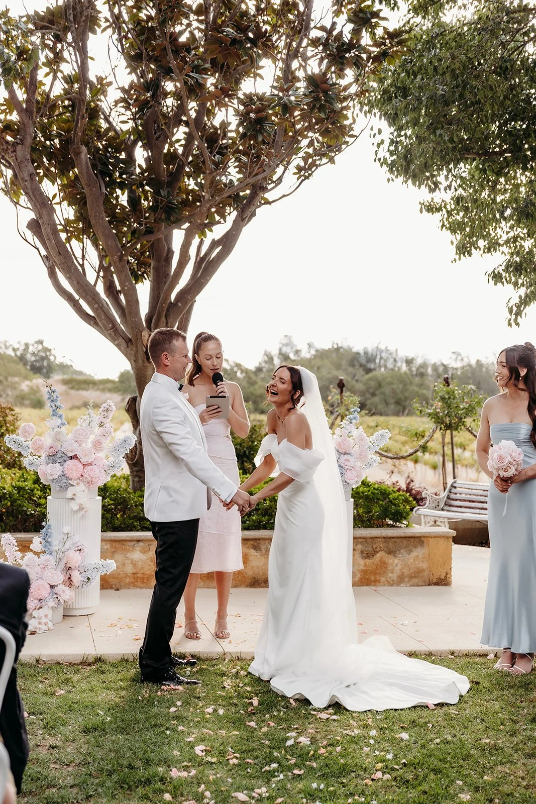 A wedding ceremony outdoors, with a bride and groom holding hands and exchanging vows, surrounded by bridesmaids and decorated with floral arrangements.