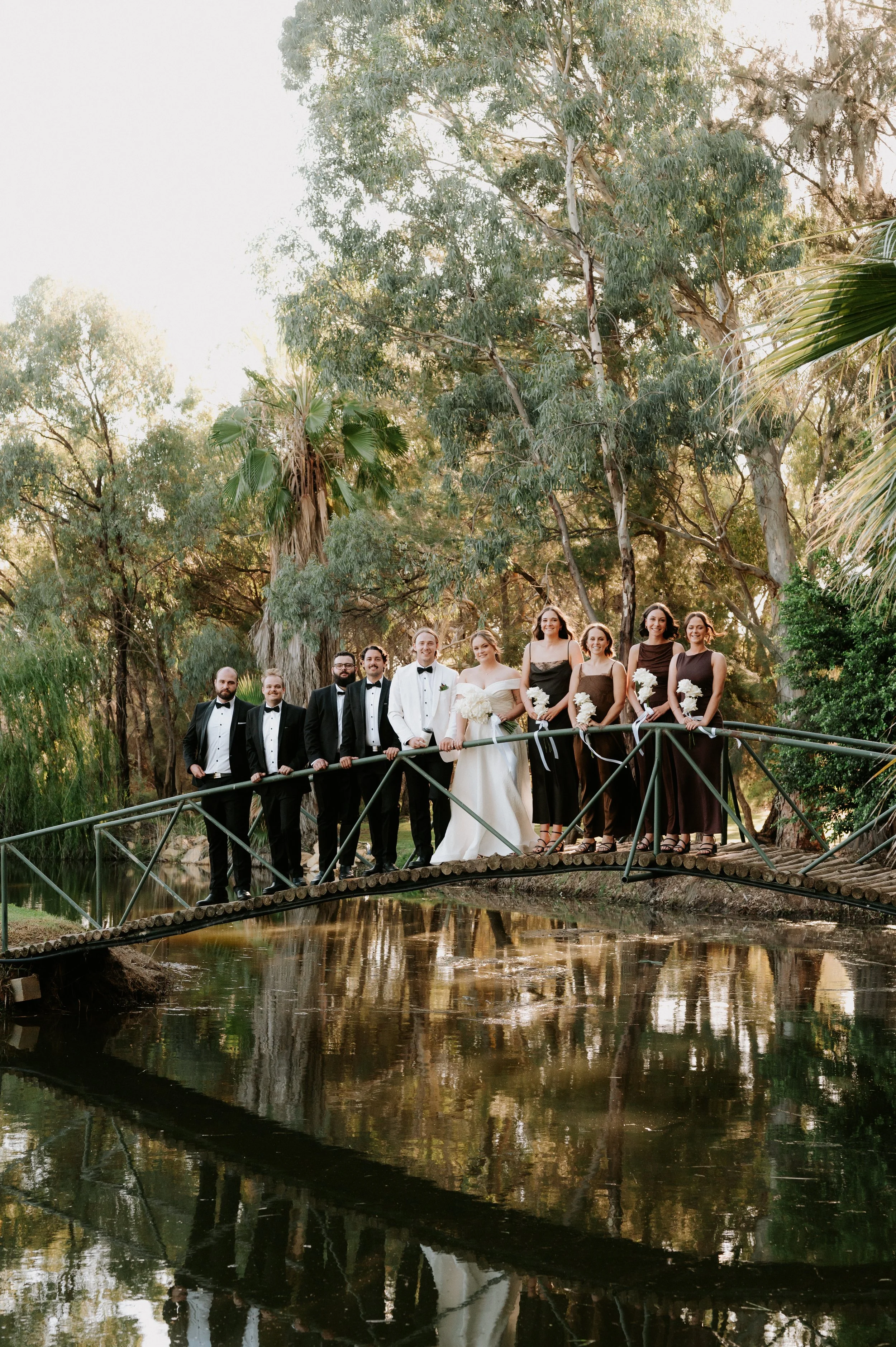 Group of people in formal attire standing on a small bridge over water, surrounded by trees.
