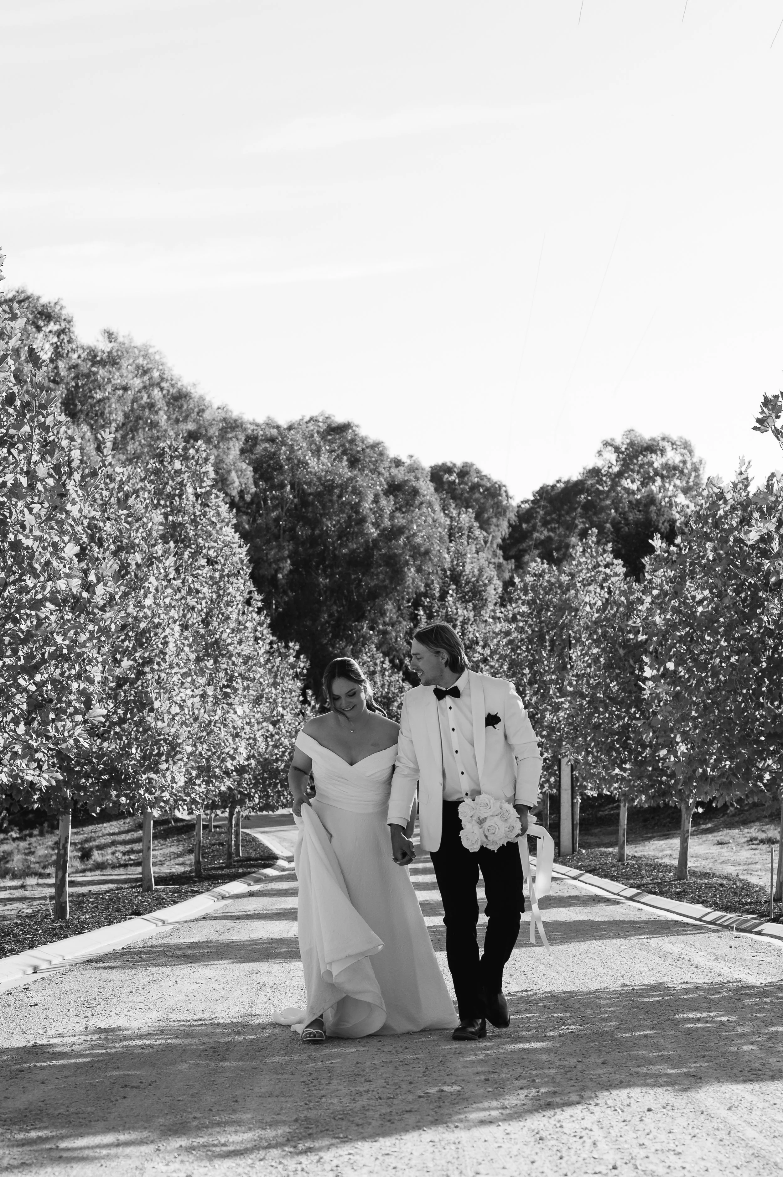 A black and white photo of a bride and groom walking hand in hand on a path lined with trees, in wedding attire, smiling and looking at each other.
